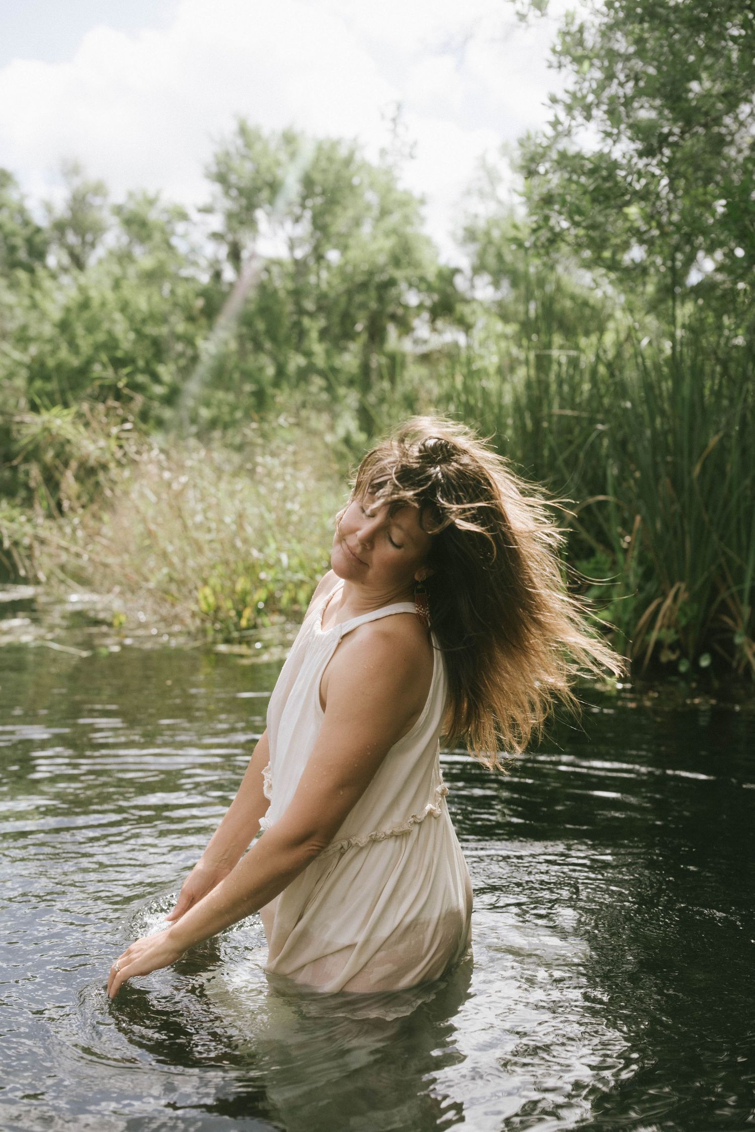 Person in white dress splashing in a serene pond surrounded by lush greenery on a summer day.