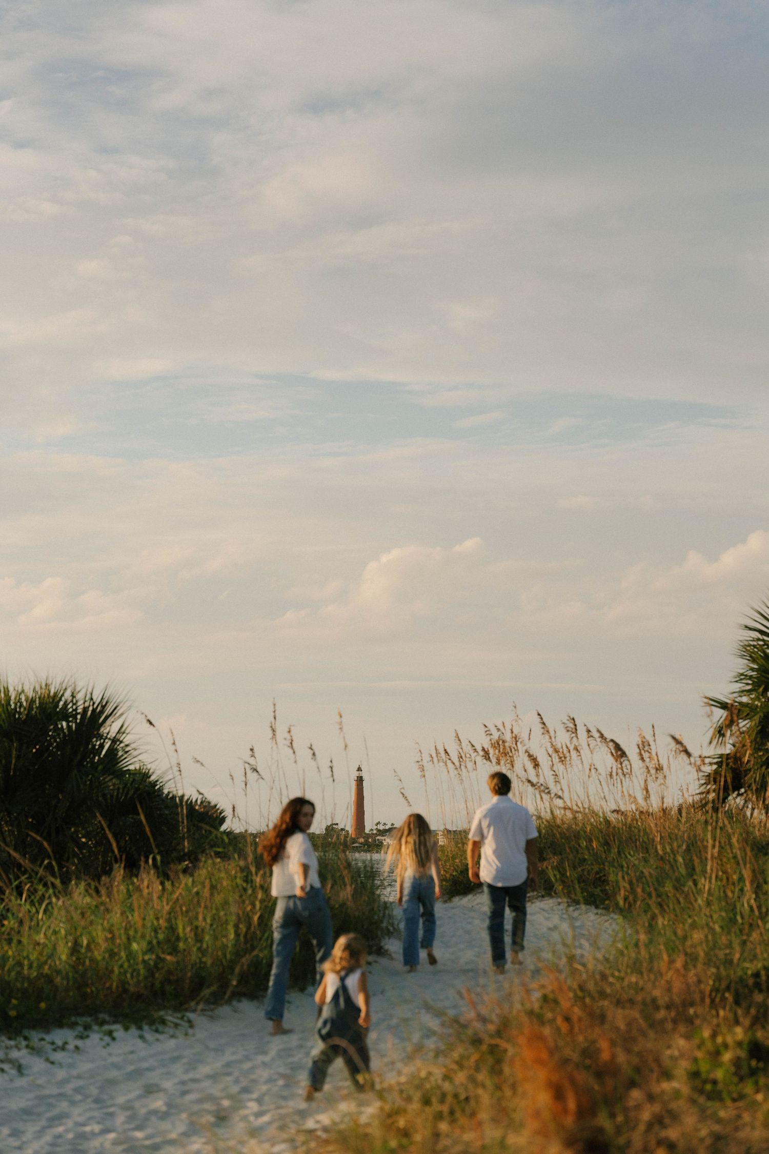 People walking along a sandy beach path through sea oats and dunes at sunset with cloudy skies.