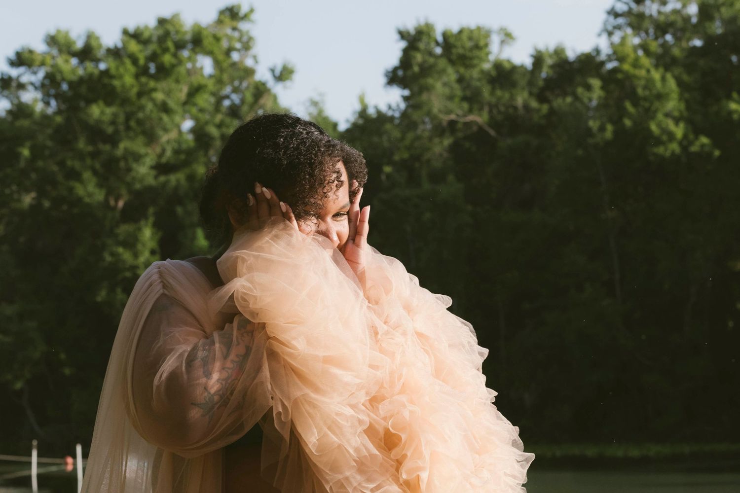 A figure in a flowing peach-colored tulle dress poses against a backdrop of green trees in natural sunlight.