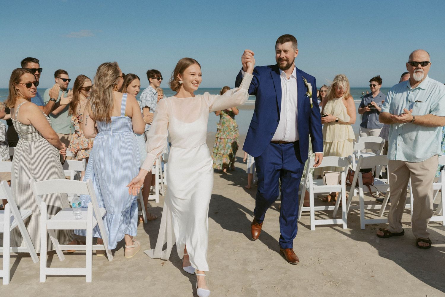 Wedding guests seated in white chairs on a beach during an outdoor ceremony with ocean views in the background.