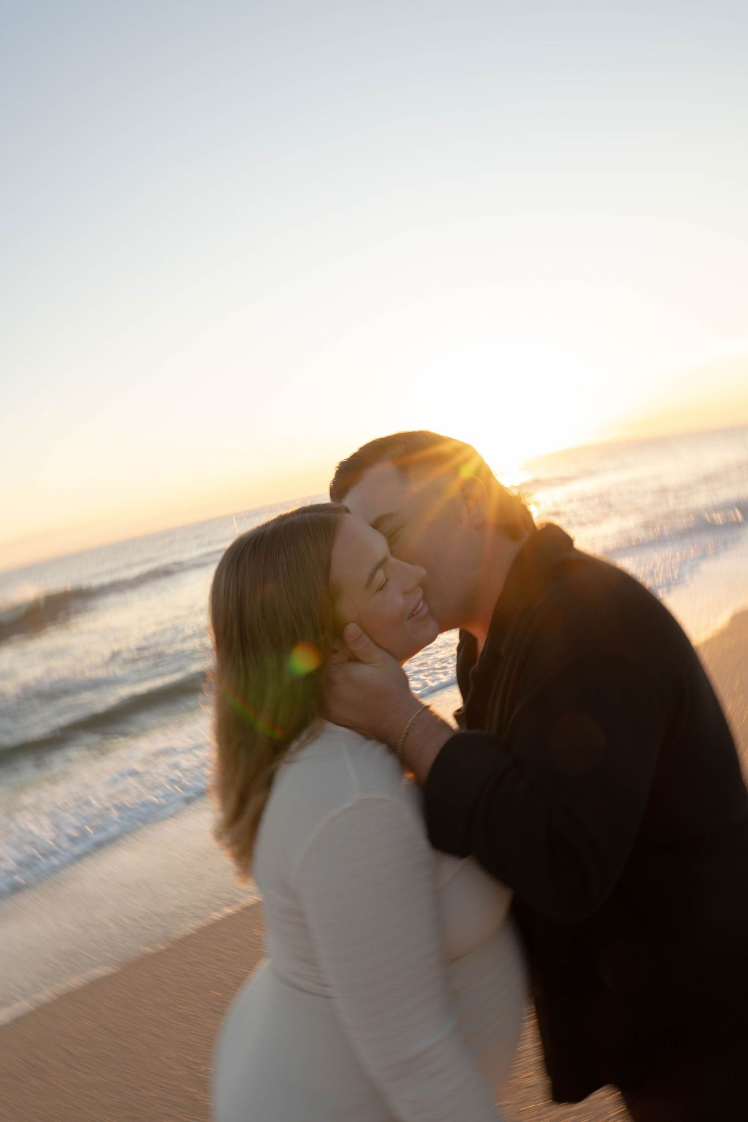 Couple sharing a romantic kiss on the beach at golden sunset, waves crashing behind them in warm evening light.