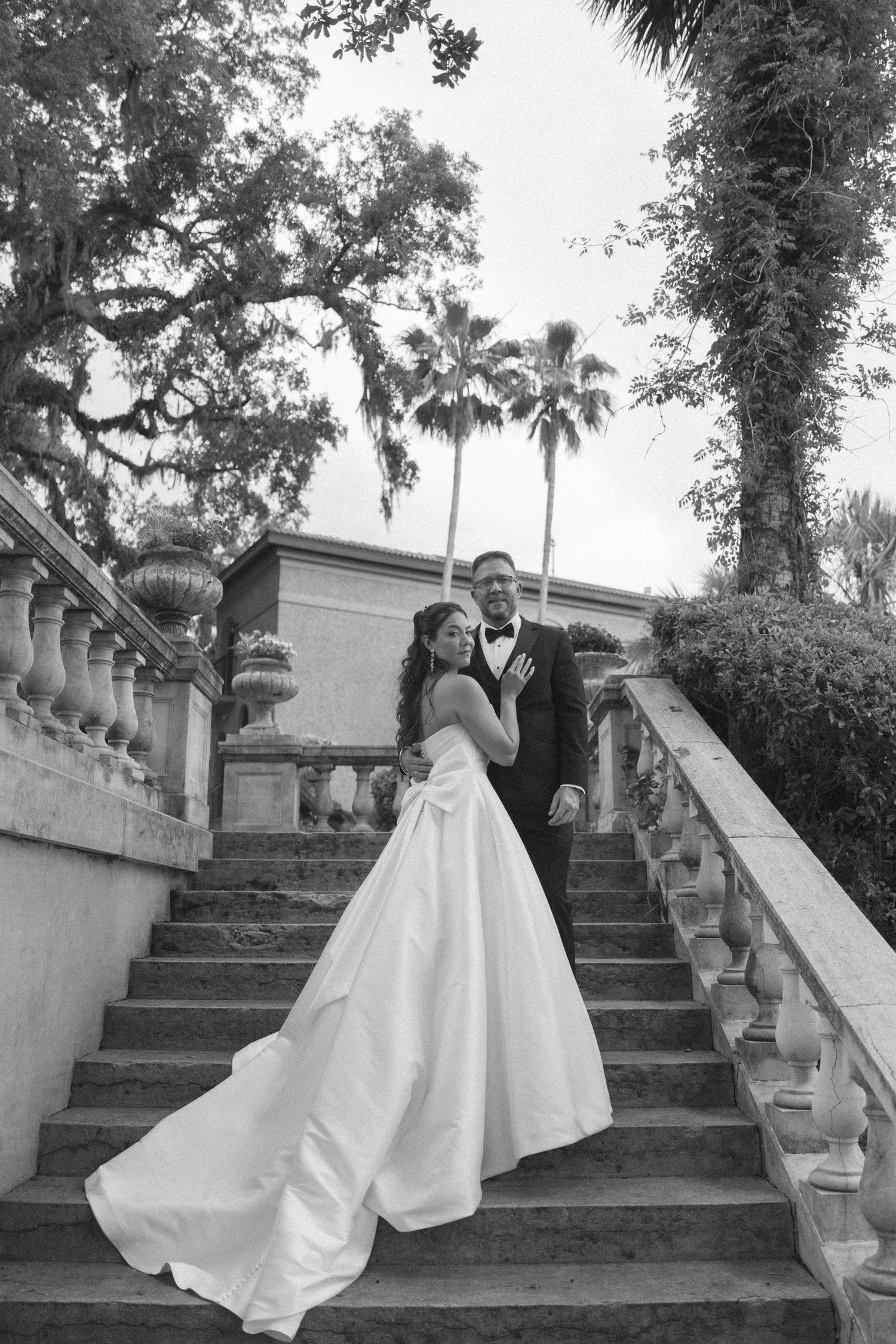 A romantic black and white wedding photo capturing a couple posing on a grand stone staircase with palm trees in the background.