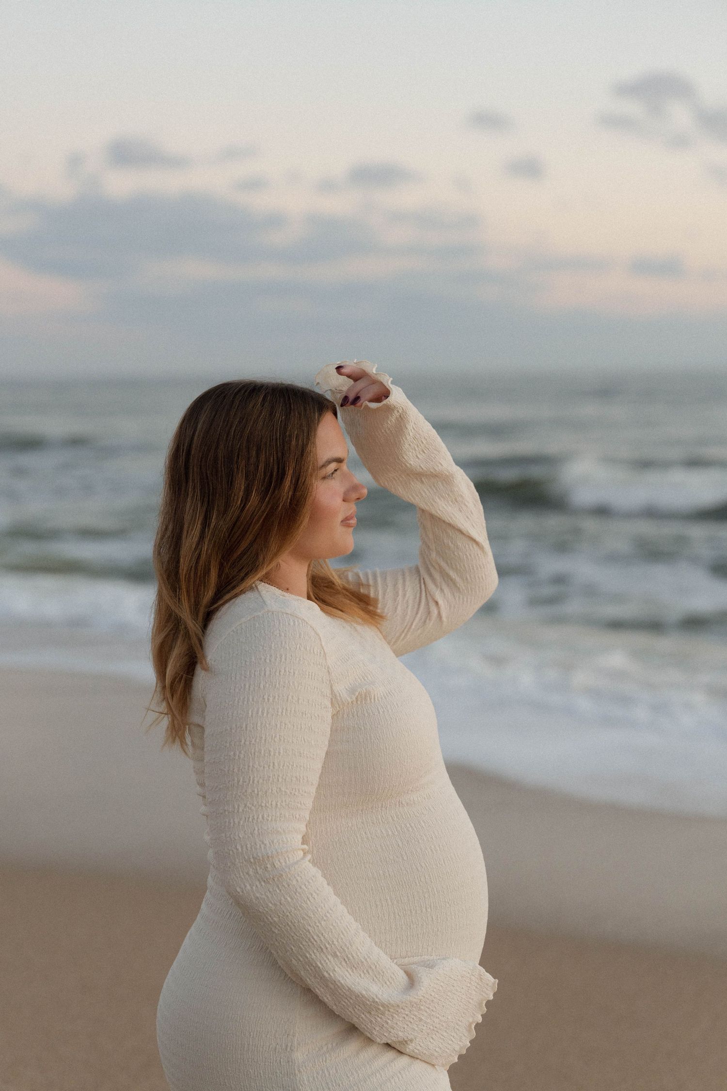 Pregnant woman in cream knit dress standing on beach at sunset, hand raised to forehead, gazing at ocean waves.