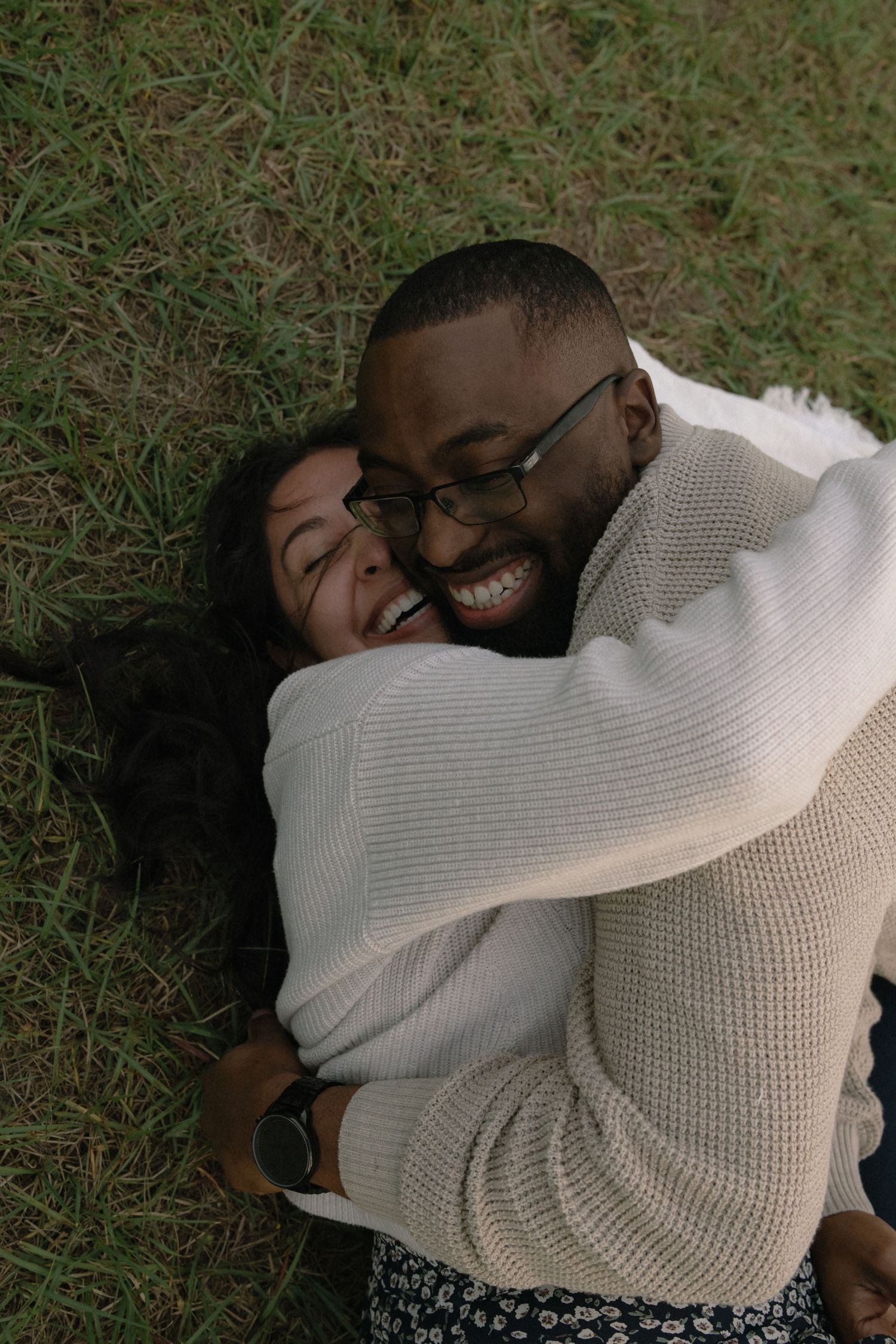 A couple in cozy cream sweaters sharing an intimate embrace while lying on grass outdoors.