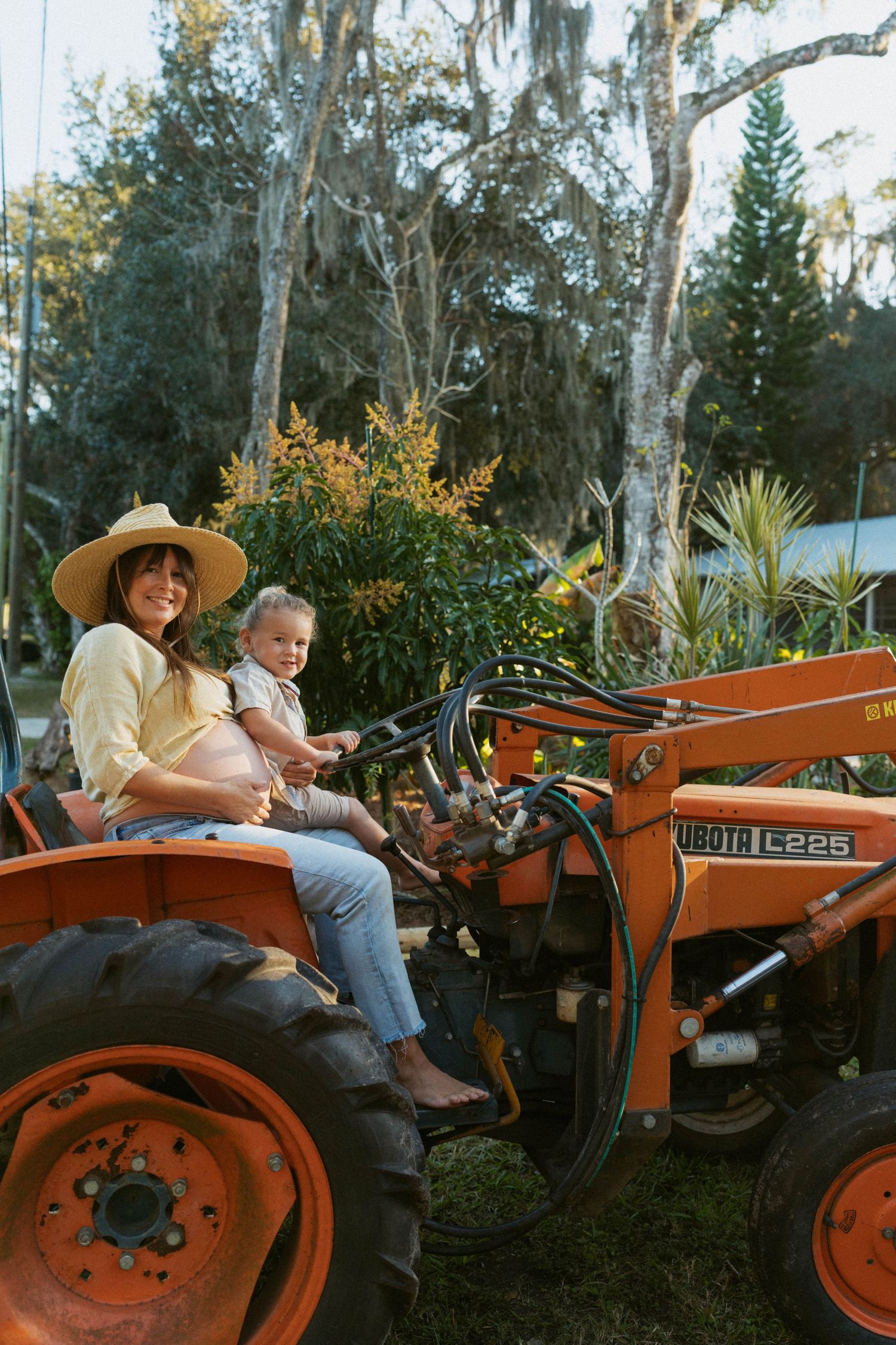 Two people sit together on an orange vintage tractor in a lush garden setting at dusk.