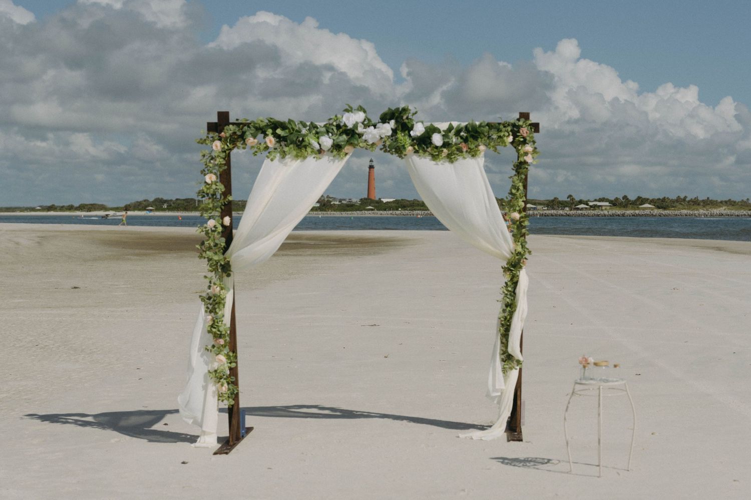 Simple beach wedding arch decorated with white fabric and greenery stands against blue ocean and sky.