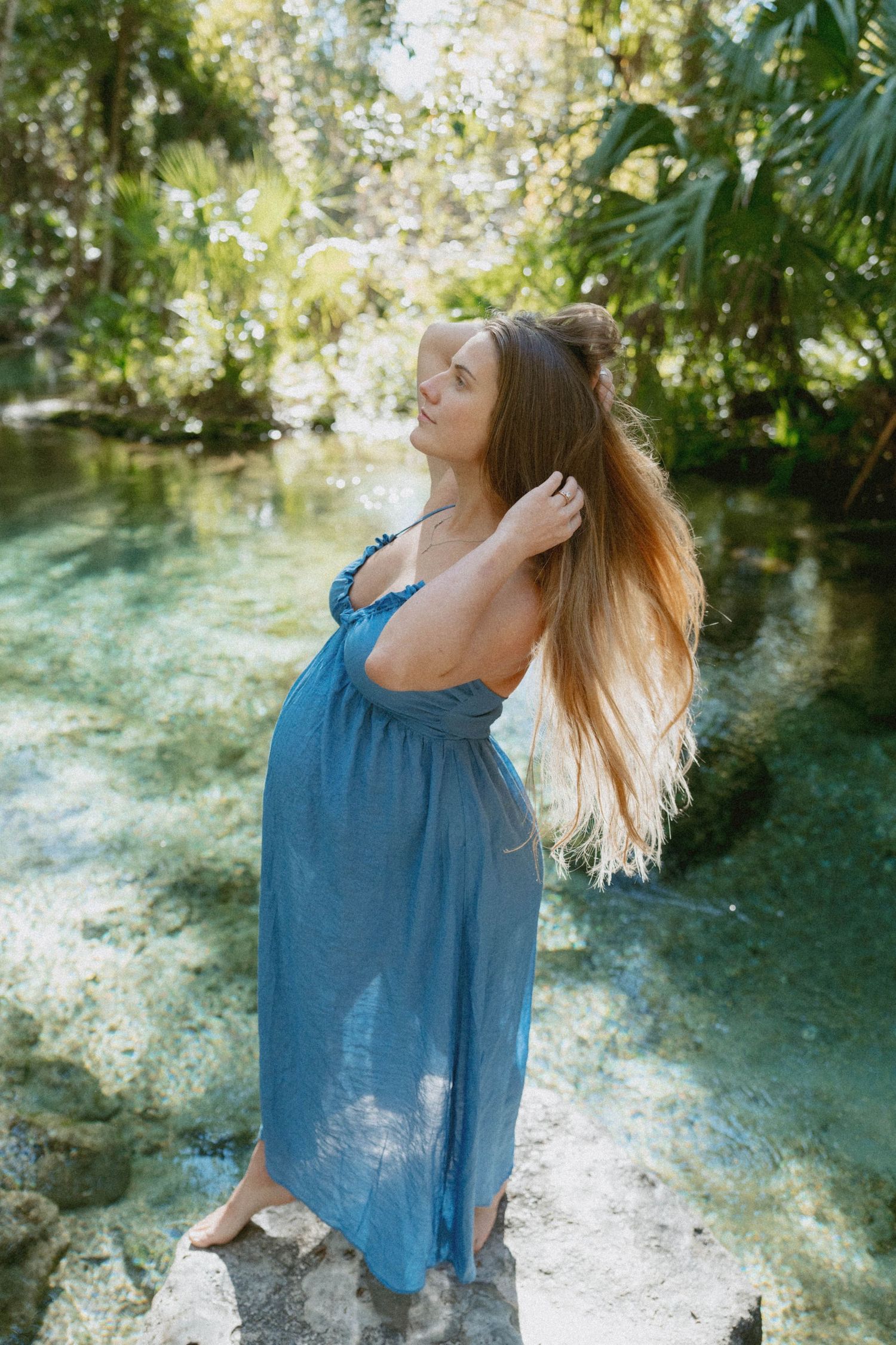 A woman in a flowing blue top dances freely in a sunlit garden setting with natural foliage in the background.