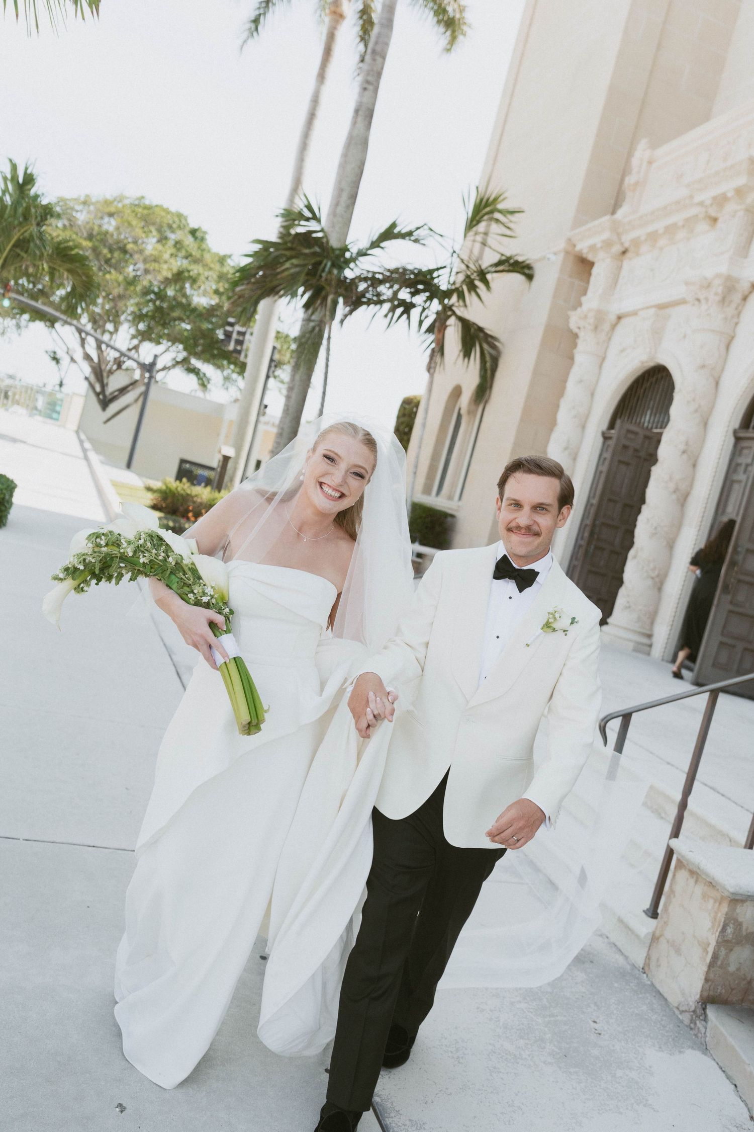 Bride in white strapless gown and groom in white tuxedo jacket hold hands smiling outside a church with palm trees.