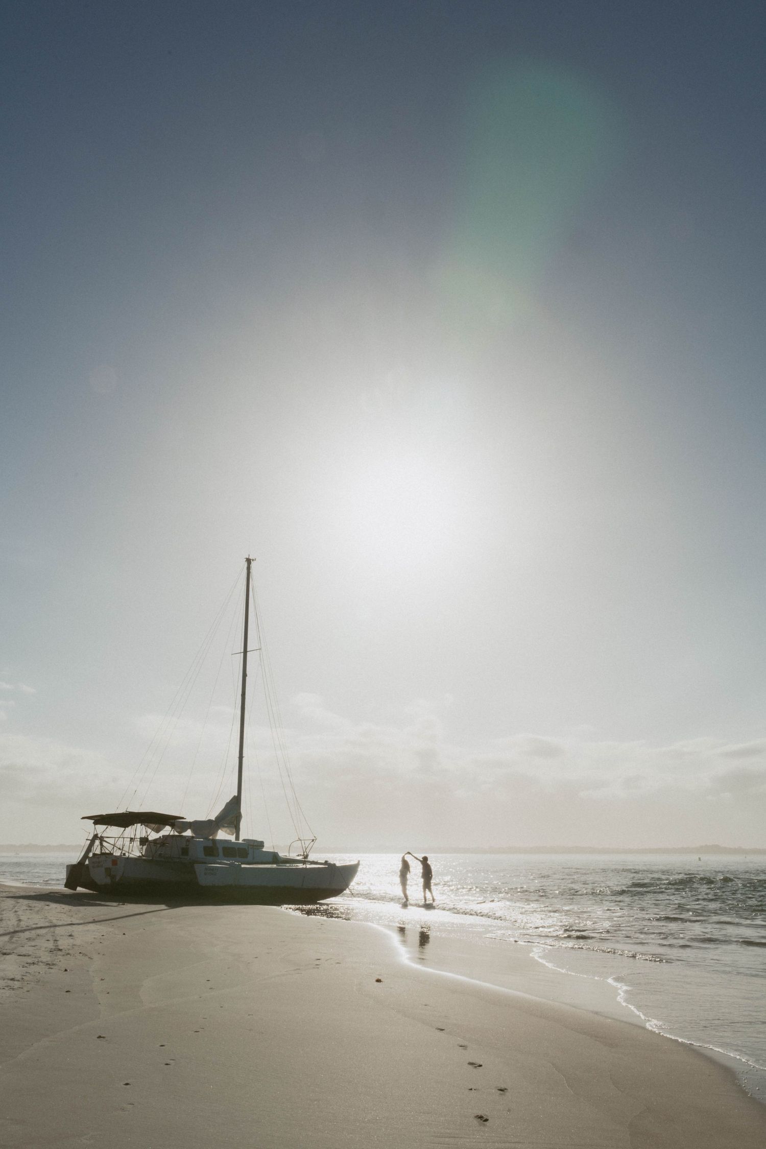 Silhouette of two people standing by a sailboat on a sunny beach with bright sun and calm ocean waves.