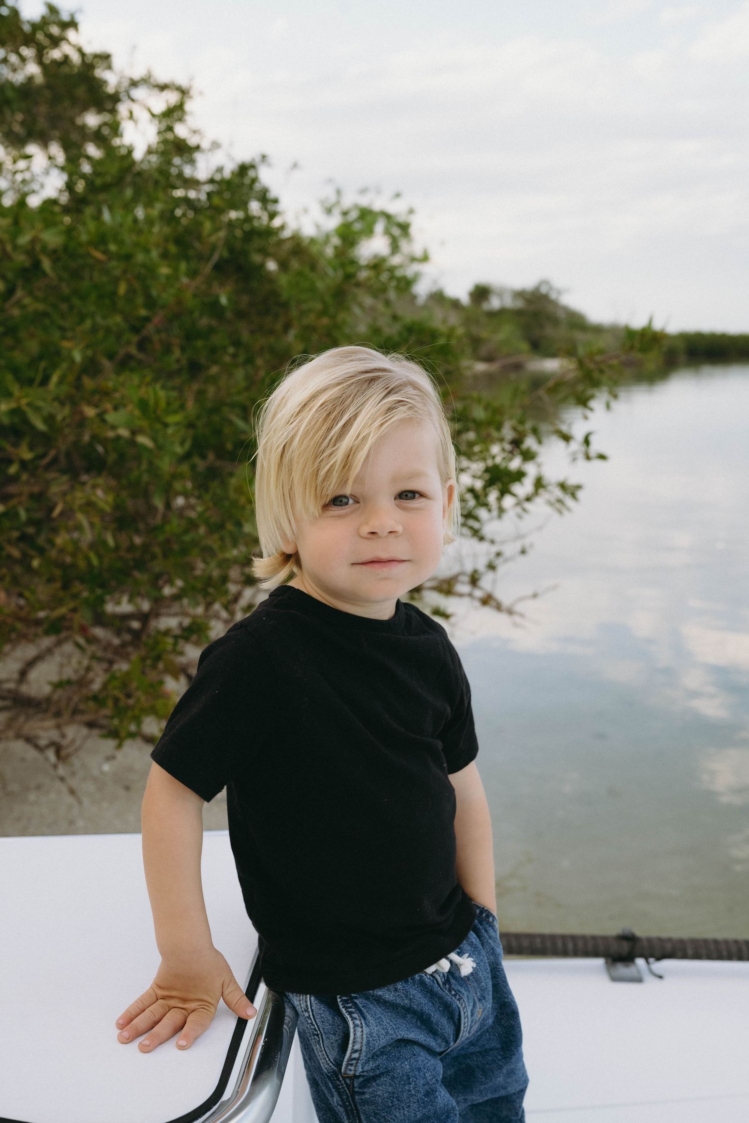 A small child in a black t-shirt and jeans poses outdoors near a calm waterway and greenery.
