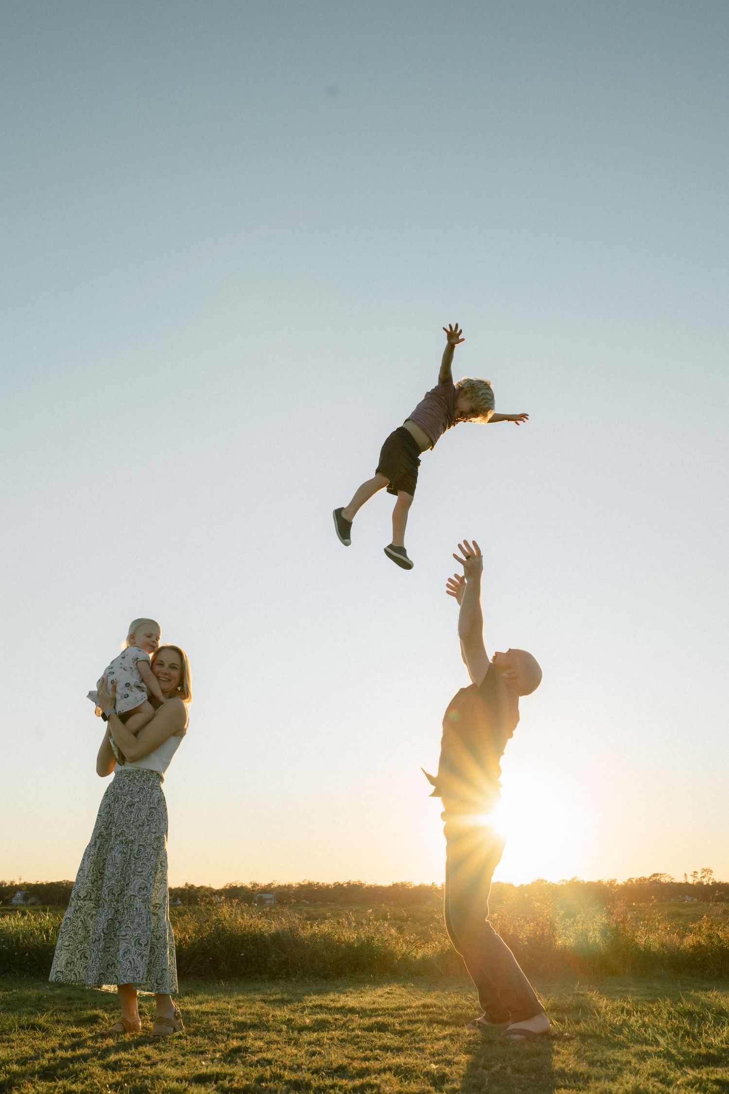 Family playing in a field at sunset with parent tossing child in the air while others watch from side.