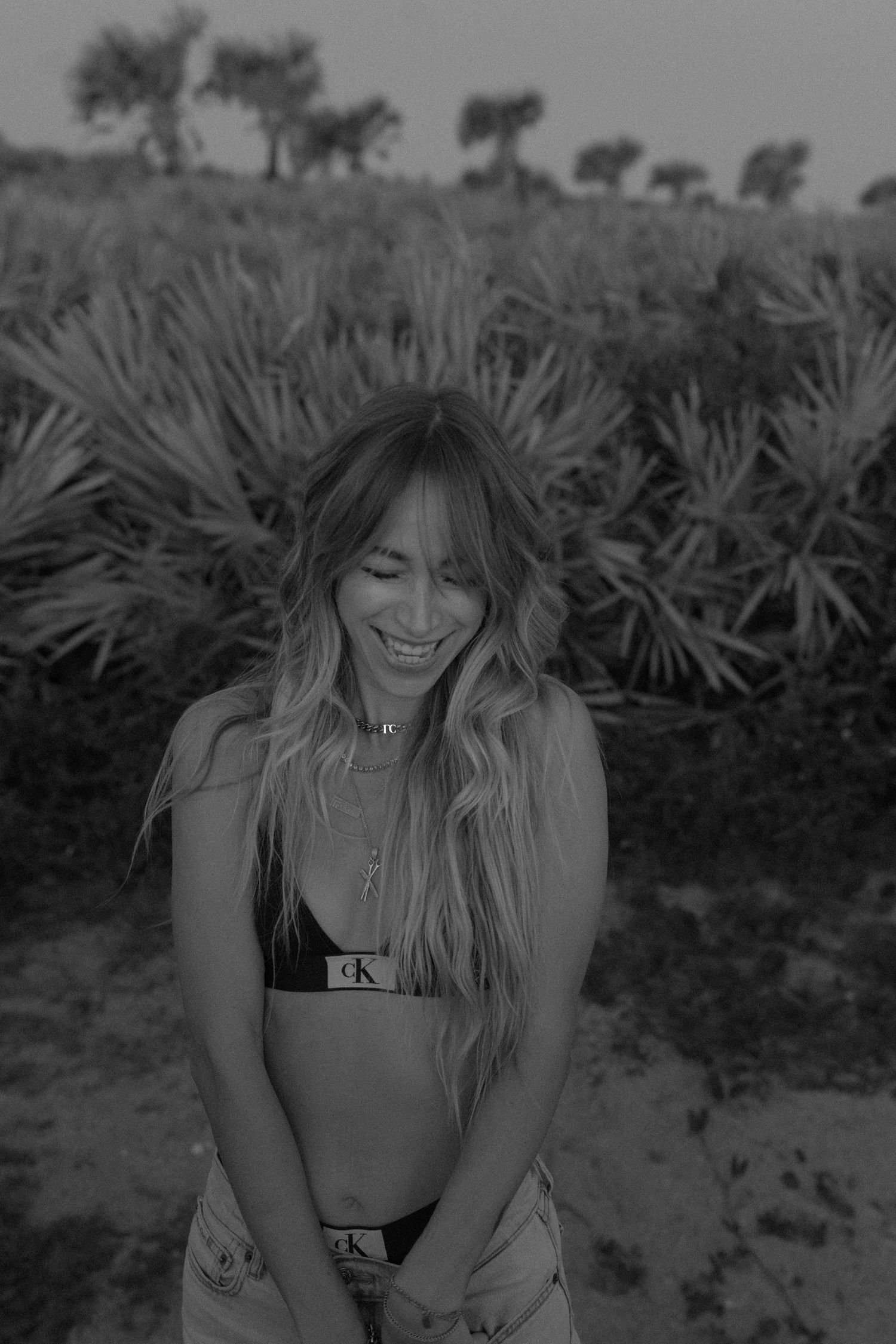 Black and white photo of someone laughing outdoors in a bikini top with palm trees visible in the background.