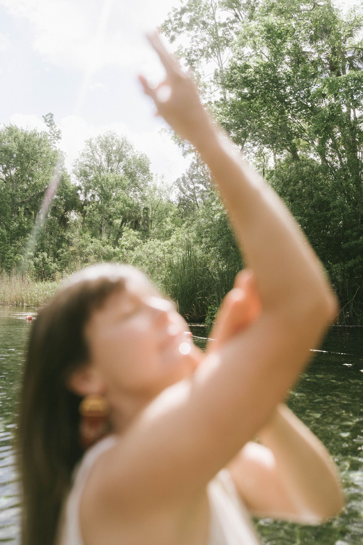 A person reaches up towards sunlight while standing near a tranquil pond surrounded by trees.