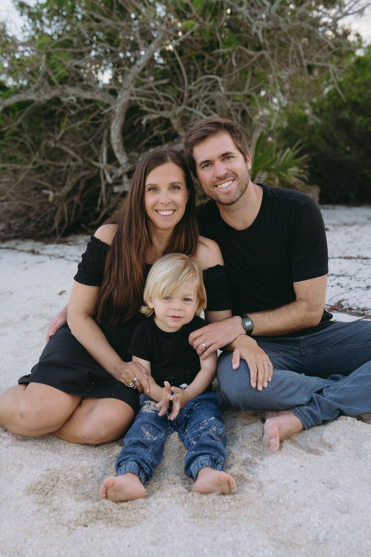 A family in matching black outfits sits together on a sandy beach with trees in the background.