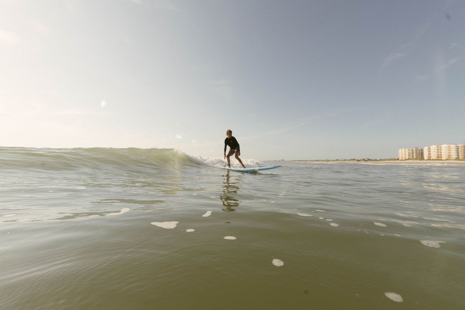 A surfer rides a gentle wave on calm waters with beachfront buildings visible in the distance on a hazy day.