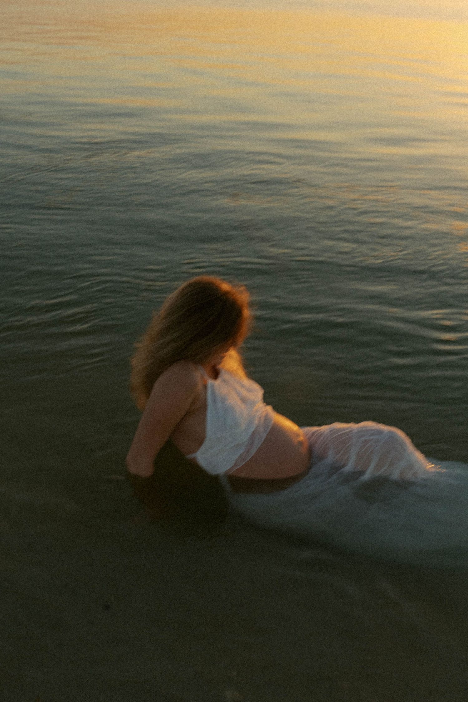 A woman in a white dress sits in shallow ocean water during a golden sunset, her long hair blowing gently.