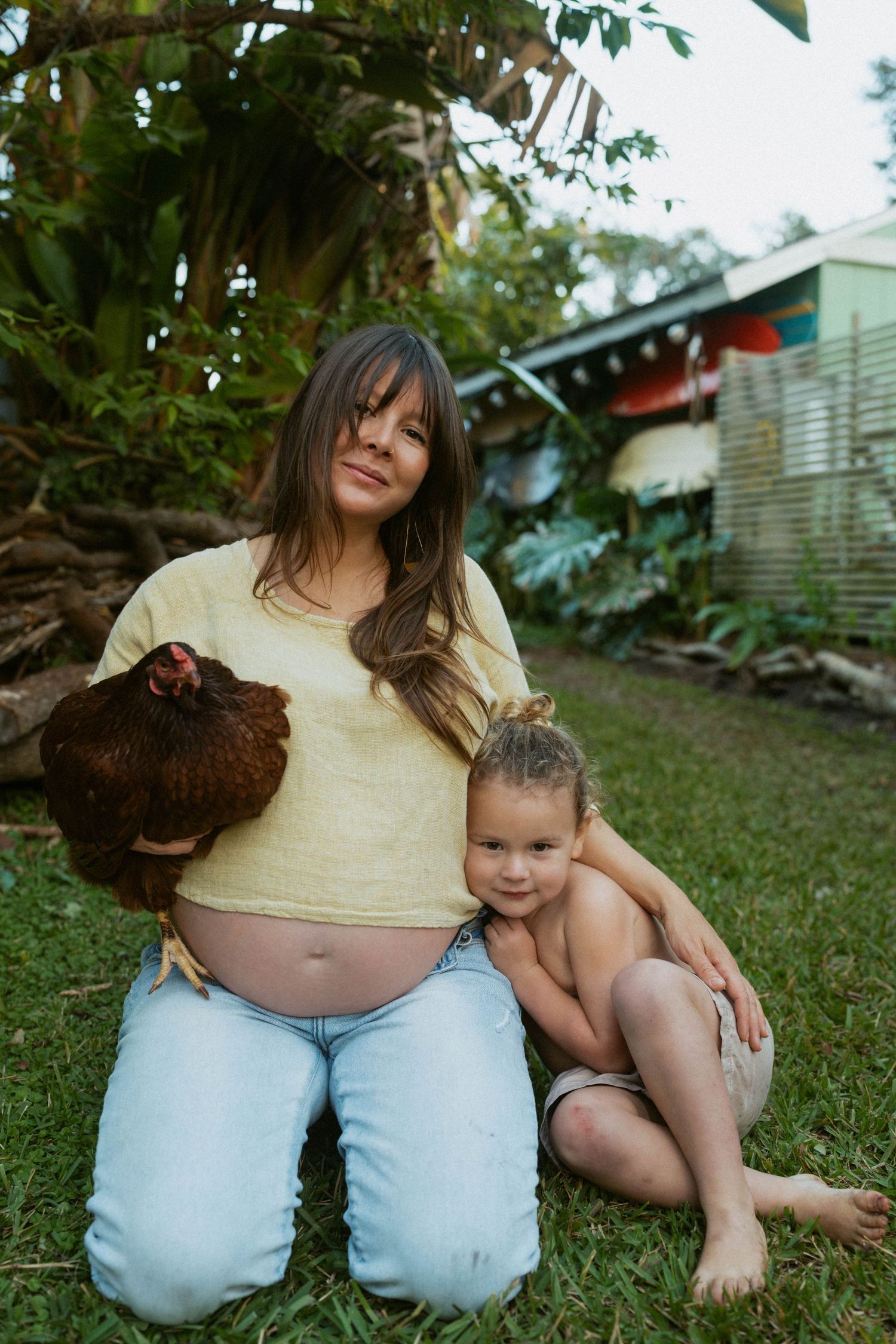 A pregnant woman in a yellow top kneels in a garden with a brown chicken and child on a sunny afternoon.
