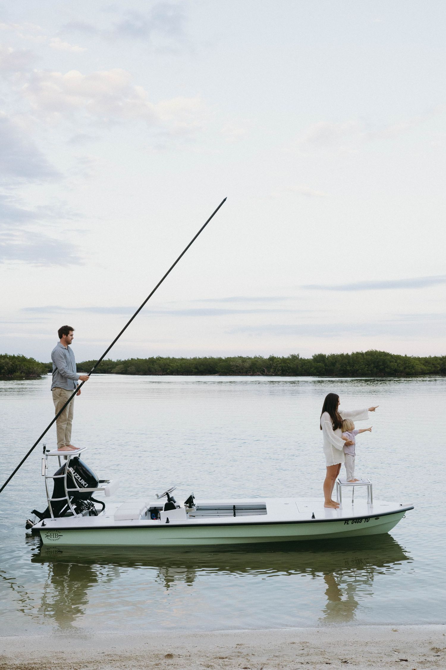 Two people fishing from a small white boat on calm waters near a sandy shoreline at dusk.