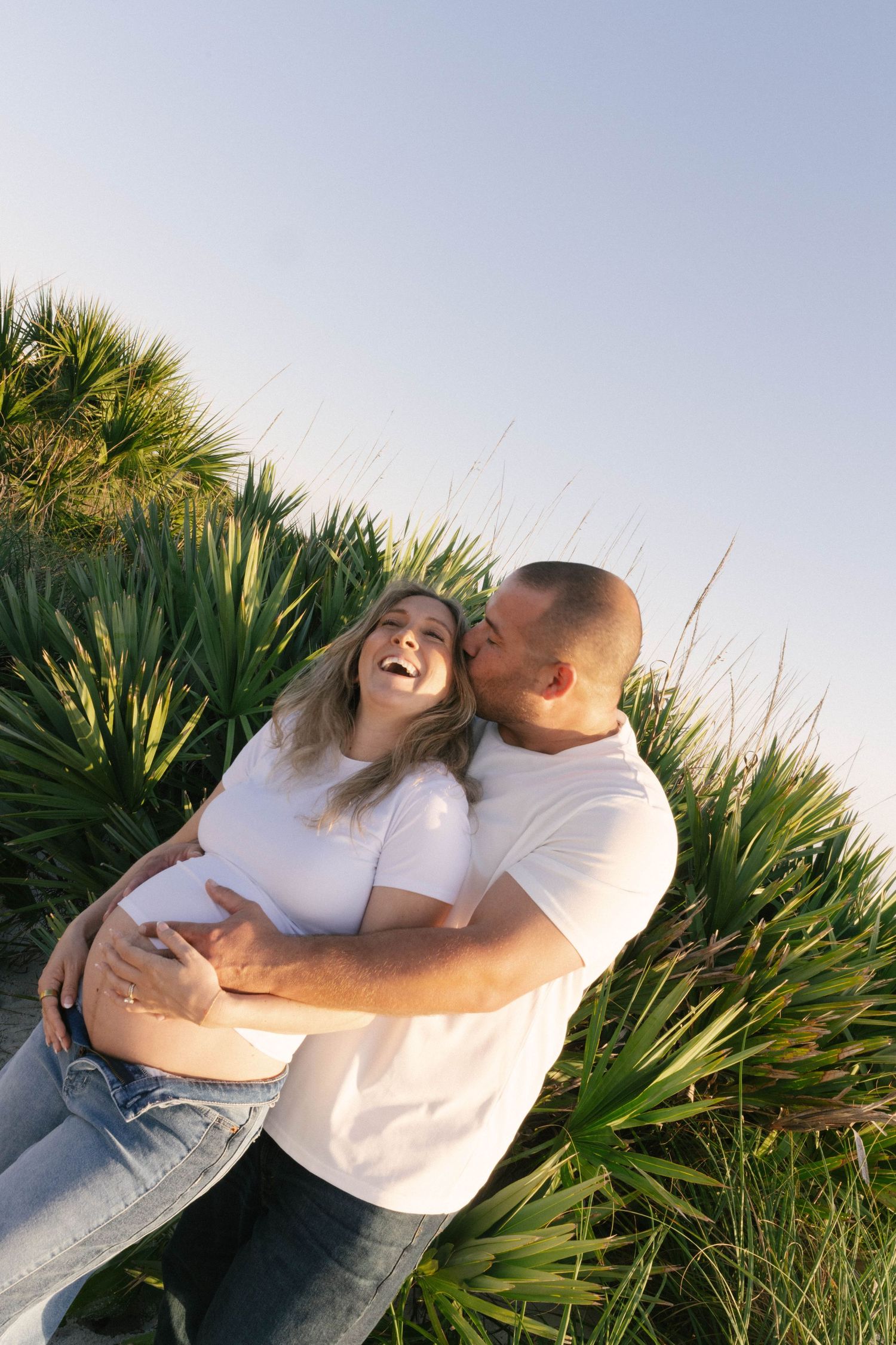 Happy pregnant couple in white shirts embracing outdoors, man kissing woman's cheek near tropical green plants.