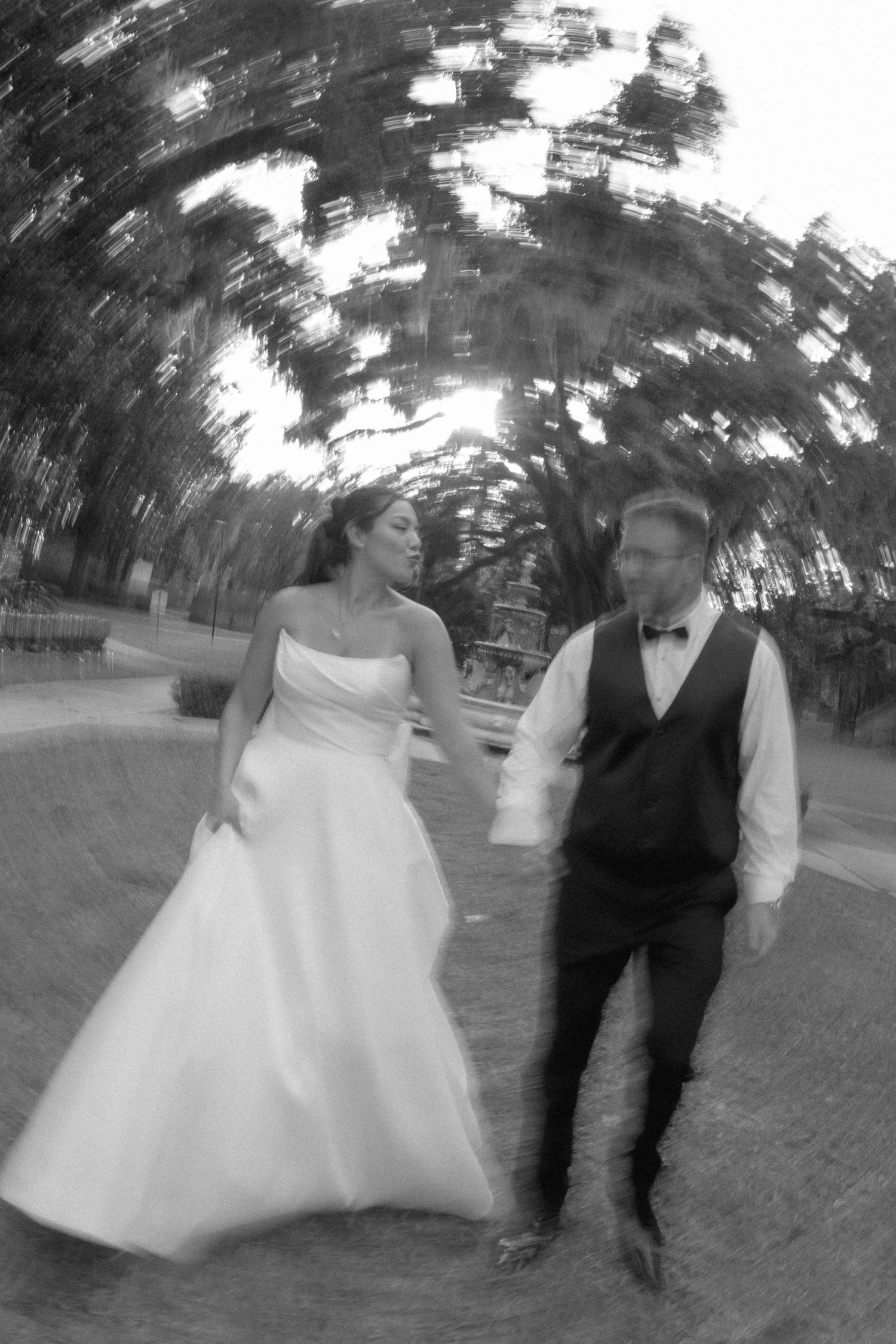 A blurred black and white artistic photo shows a couple walking together during their wedding celebration outdoors.