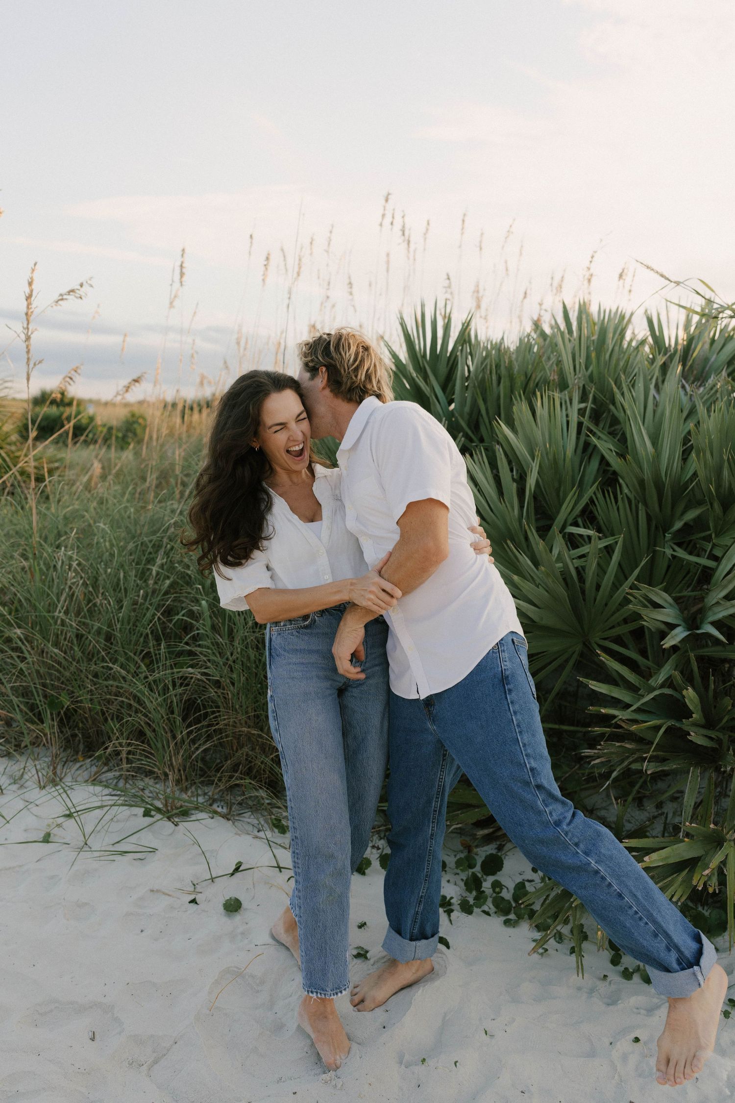 A laughing couple in white shirts and jeans embrace playfully on sandy beach dunes with green coastal grasses behind them.