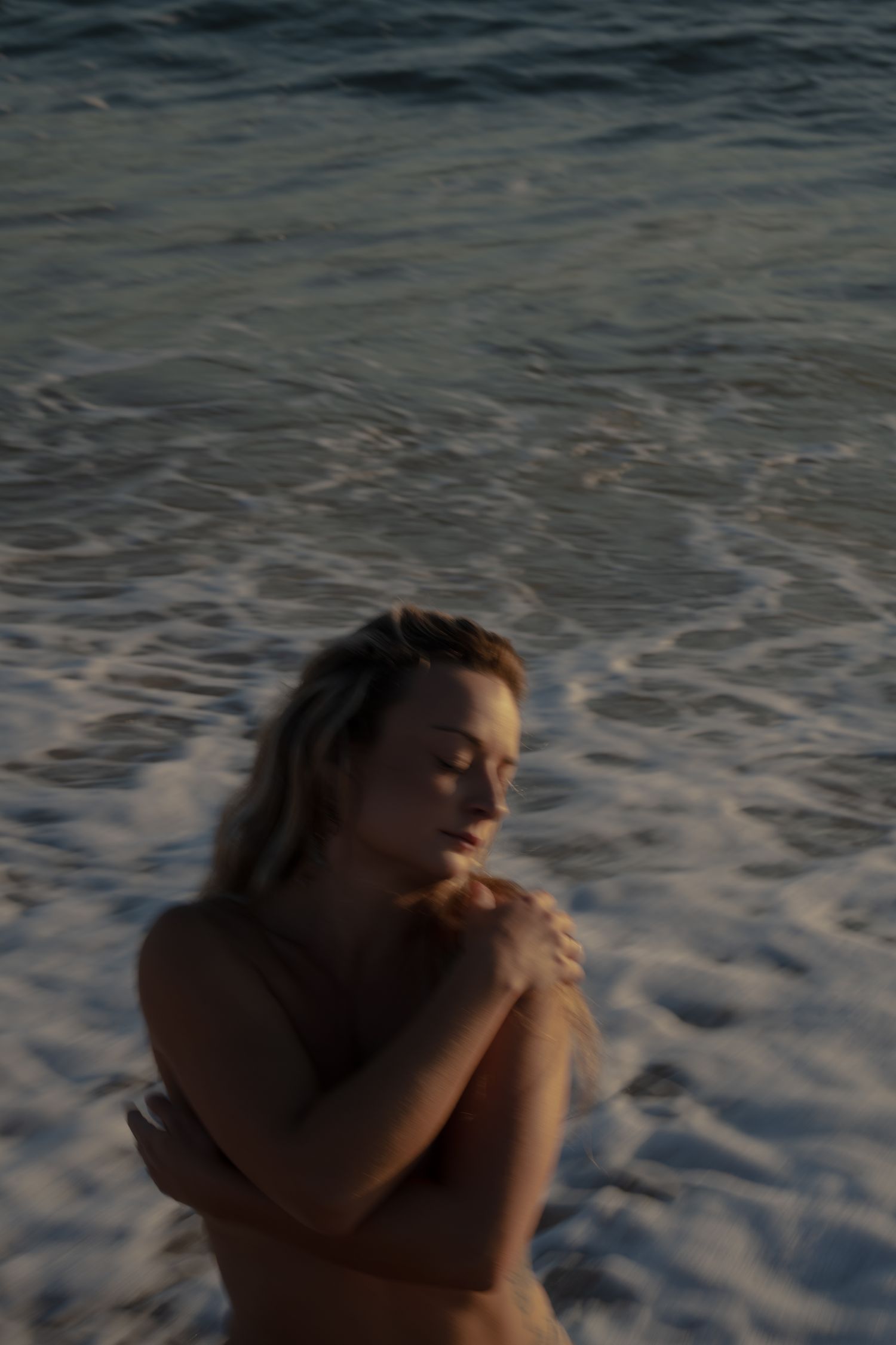 A figure stands against ocean waves at sunset with closed eyes in a dreamy, reflective pose.