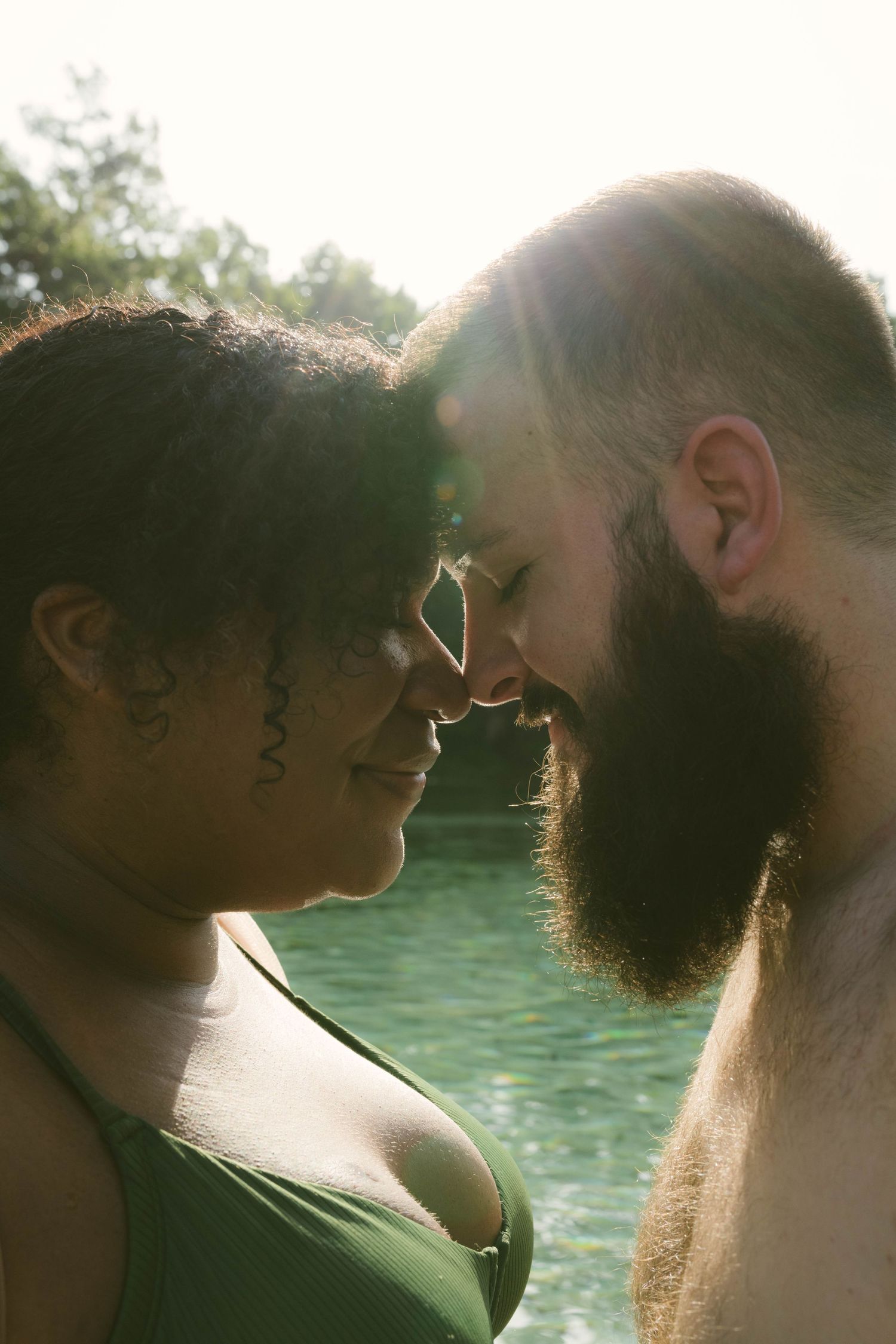 Intimate couple sharing a tender moment by the water on a sunny day with lens flare in the background.