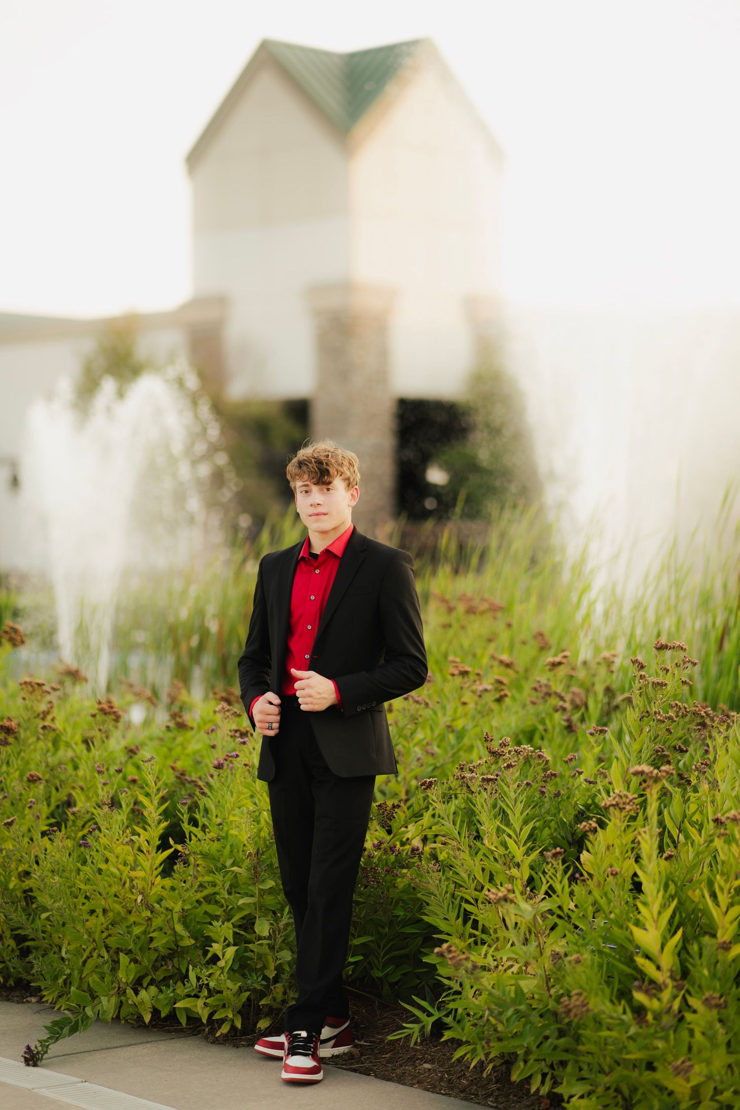 Teen boy in black suit and red shirt poses among green plants near a fountain outside a building.