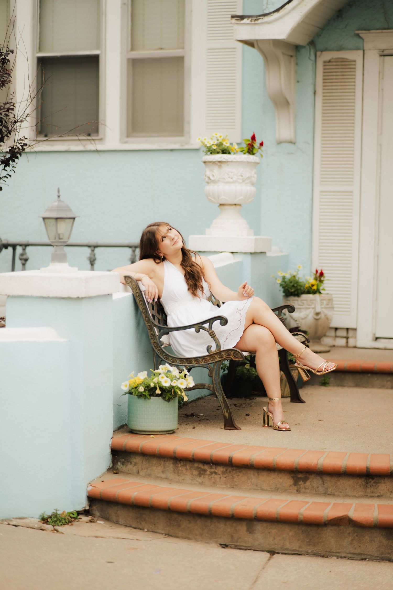 Brunette woman in white dress relaxing on iron bench outside blue Victorian house.