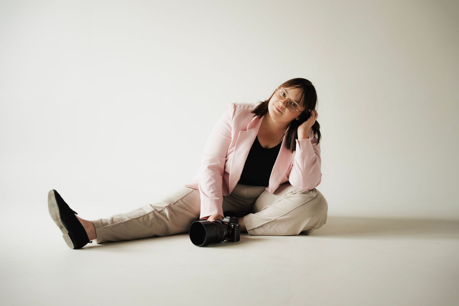 Woman in pink blazer sits on floor holding camera while posing against white background.