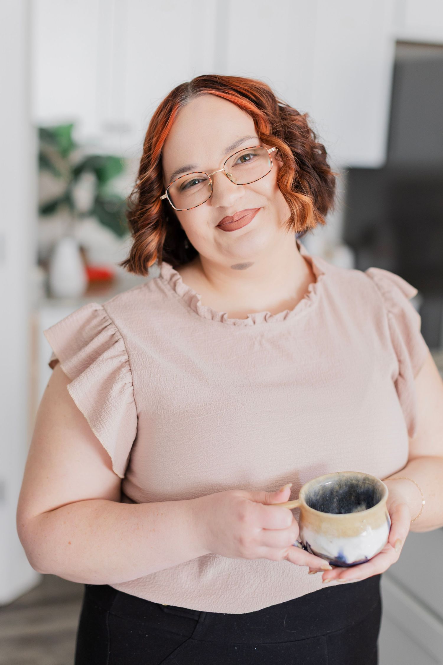 A smiling woman with red hair and glasses holds a ceramic mug in a bright modern kitchen.