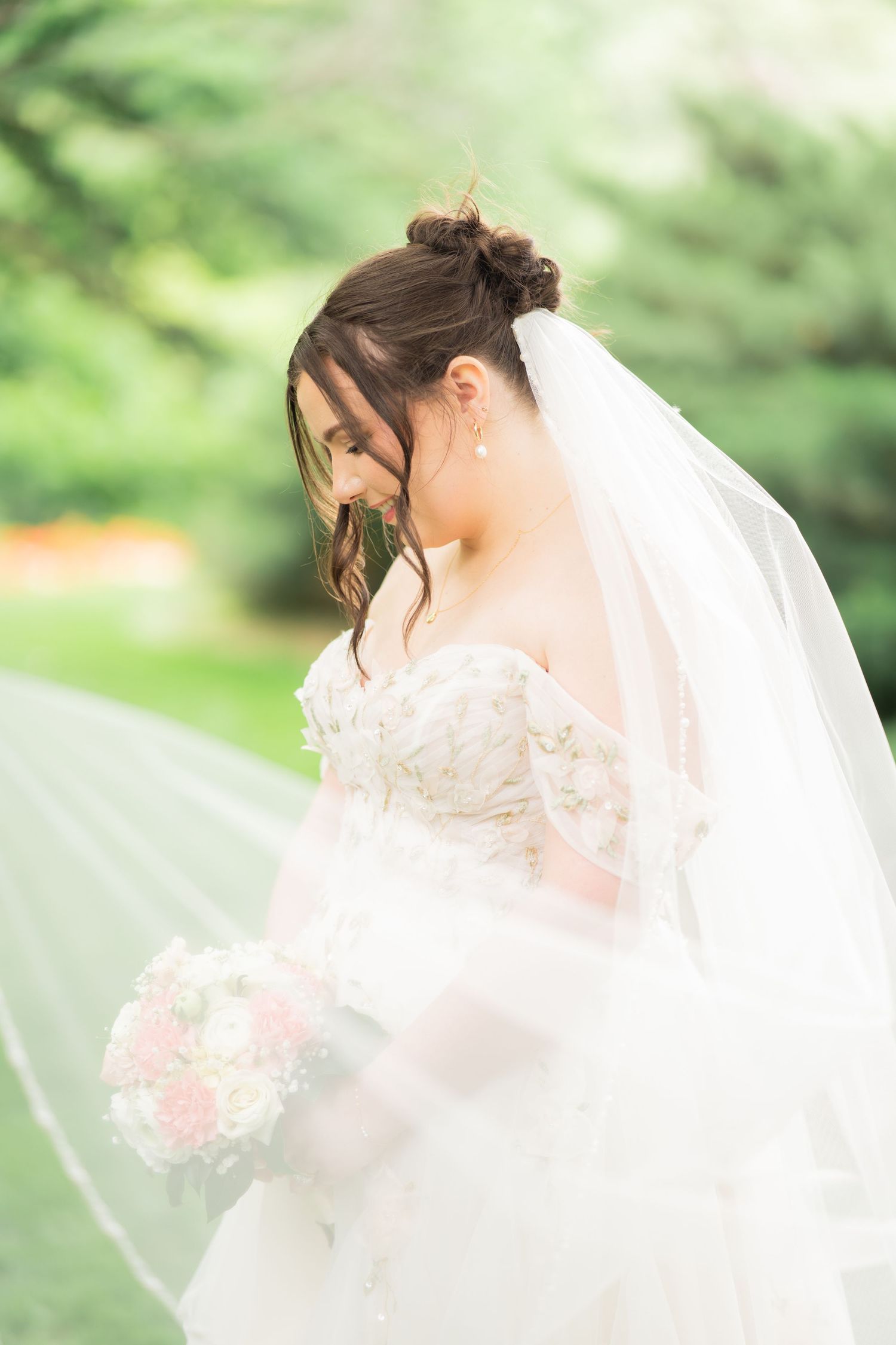 Bride in off-shoulder lace gown holds pink bouquet with veil flowing in breeze.