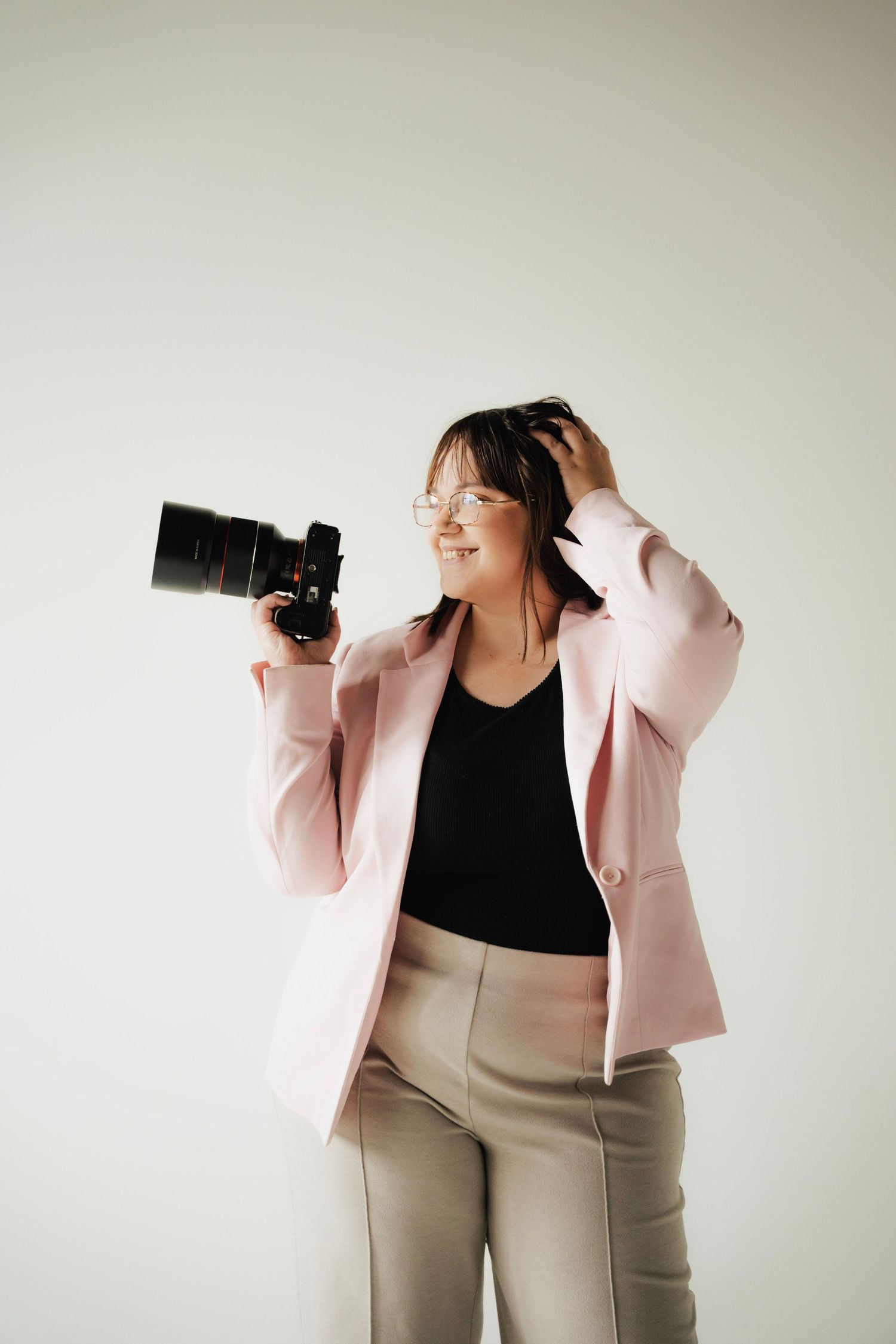 Female photographer in pink blazer holding professional camera while touching her hair indoors.