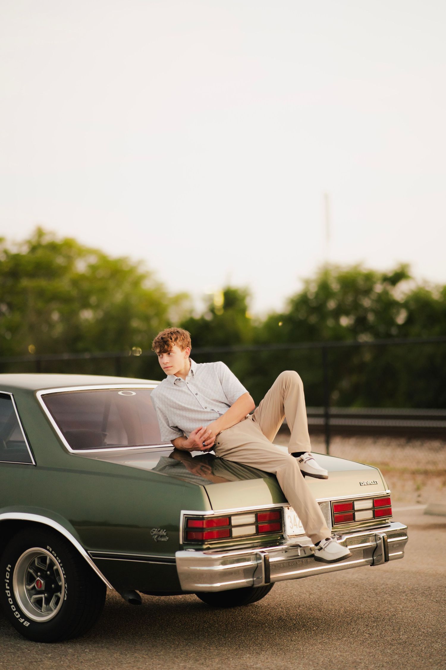 Young man in khakis sits on trunk of classic green Buick Monte Carlo at golden hour near tree-lined road.