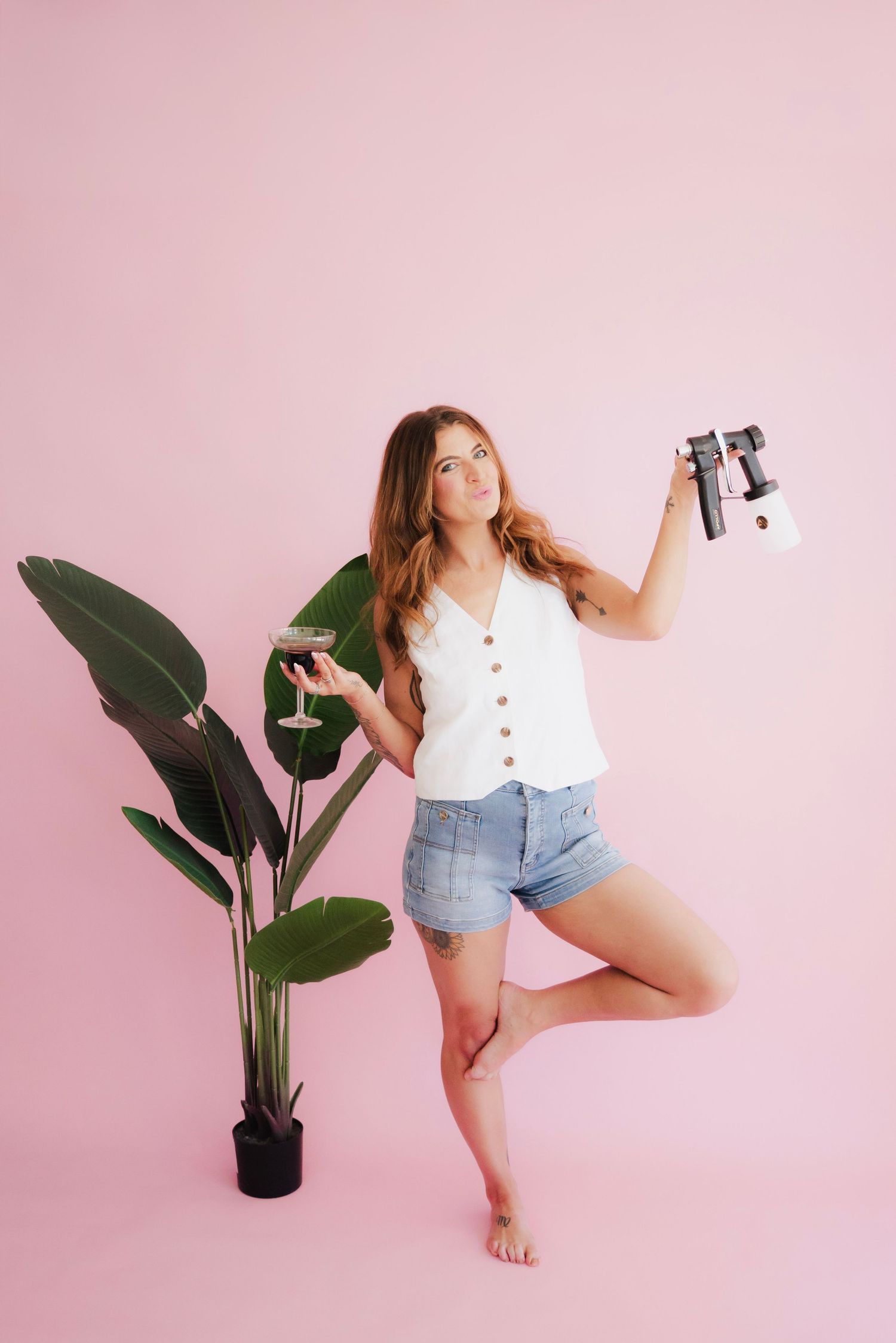 Woman in white top and denim shorts holding a camera and wine glass, posing barefoot near a tropical plant on pink background.