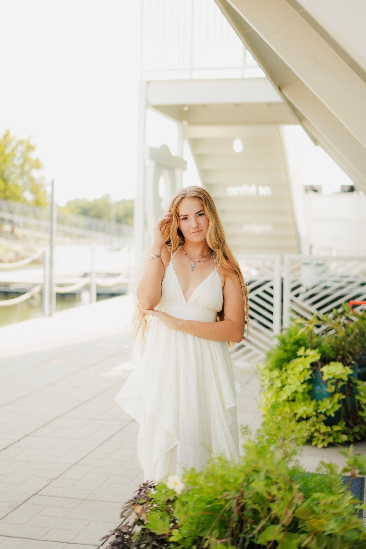 Young woman in white dress poses gracefully near a waterfront marina surrounded by lush green plants on a sunny day.