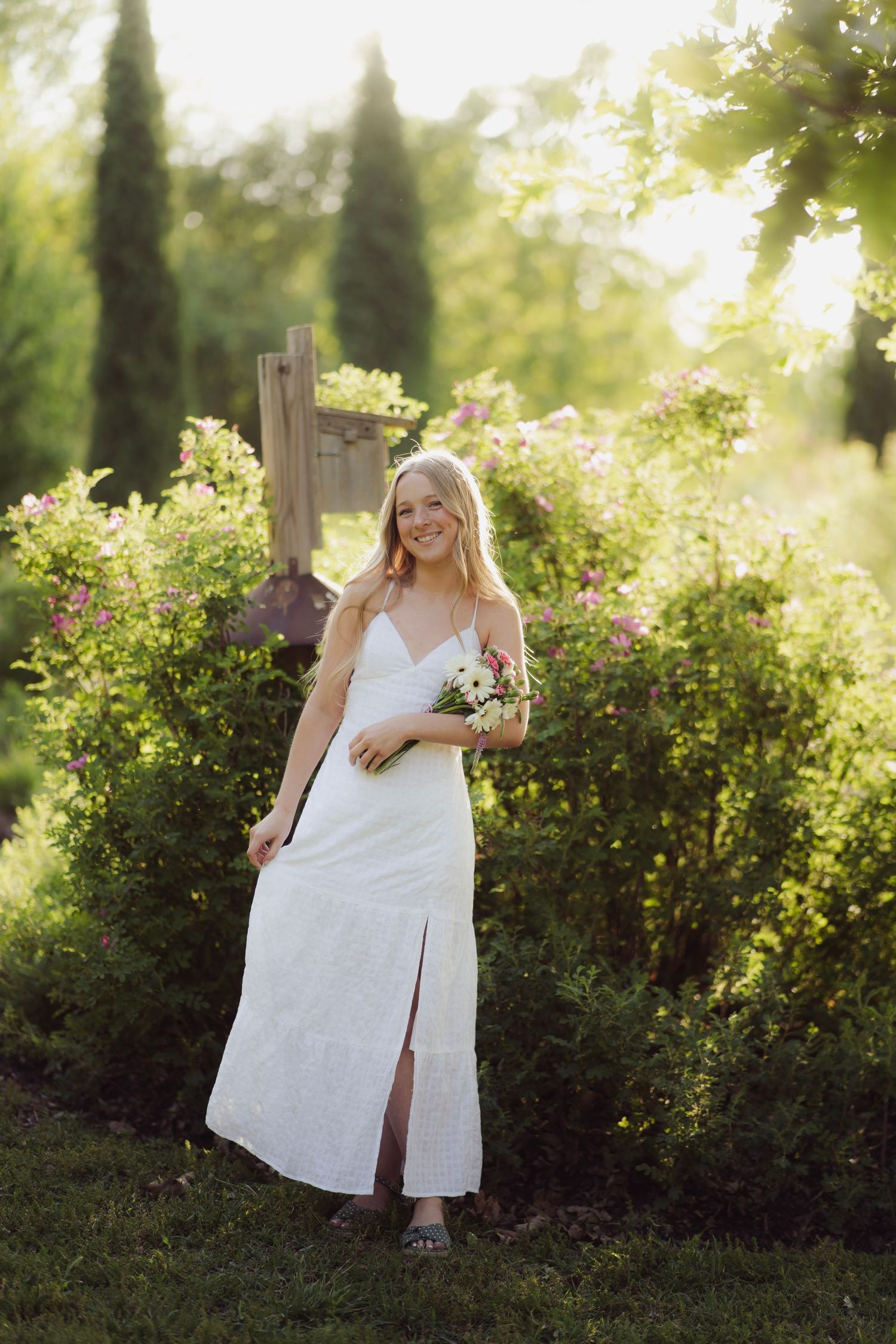 Smiling blonde woman in white maxi dress holding wildflowers in a sunlit garden.