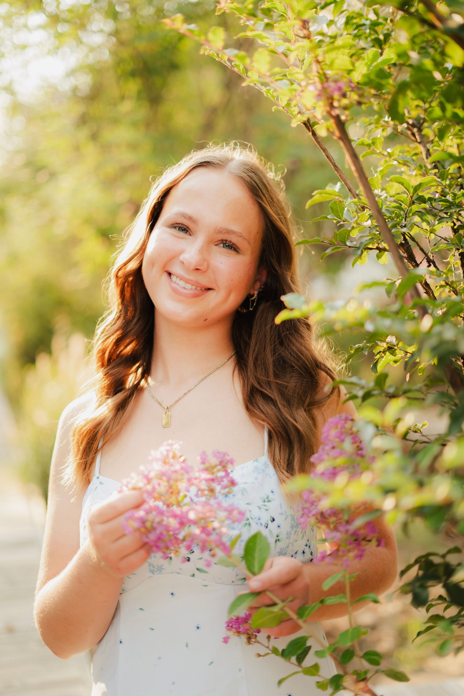Smiling young woman in white dress holding pink lilac flowers outdoors in sunlit garden setting.
