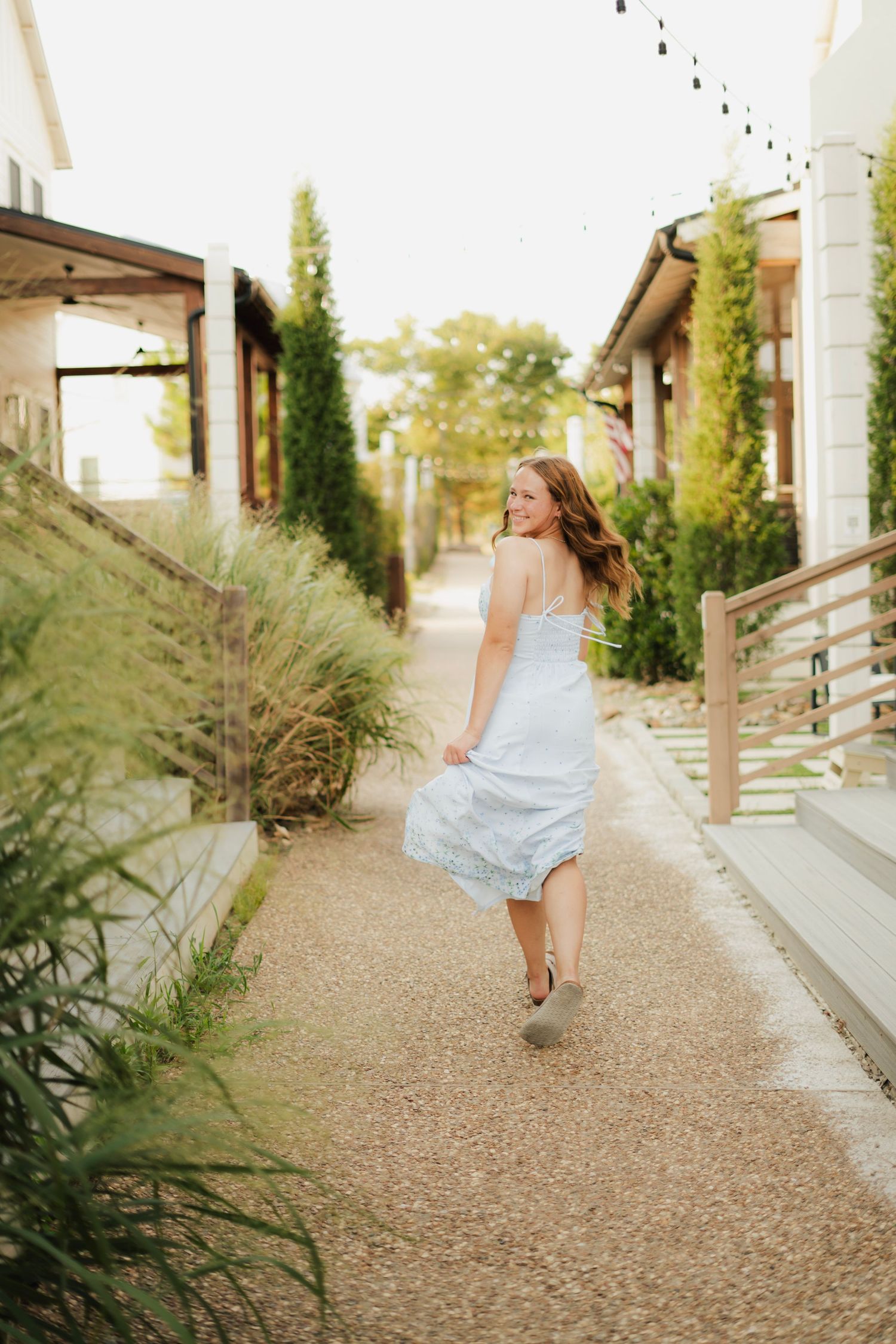 A woman in a light blue sundress walks along a charming garden pathway, glancing back with a smile.