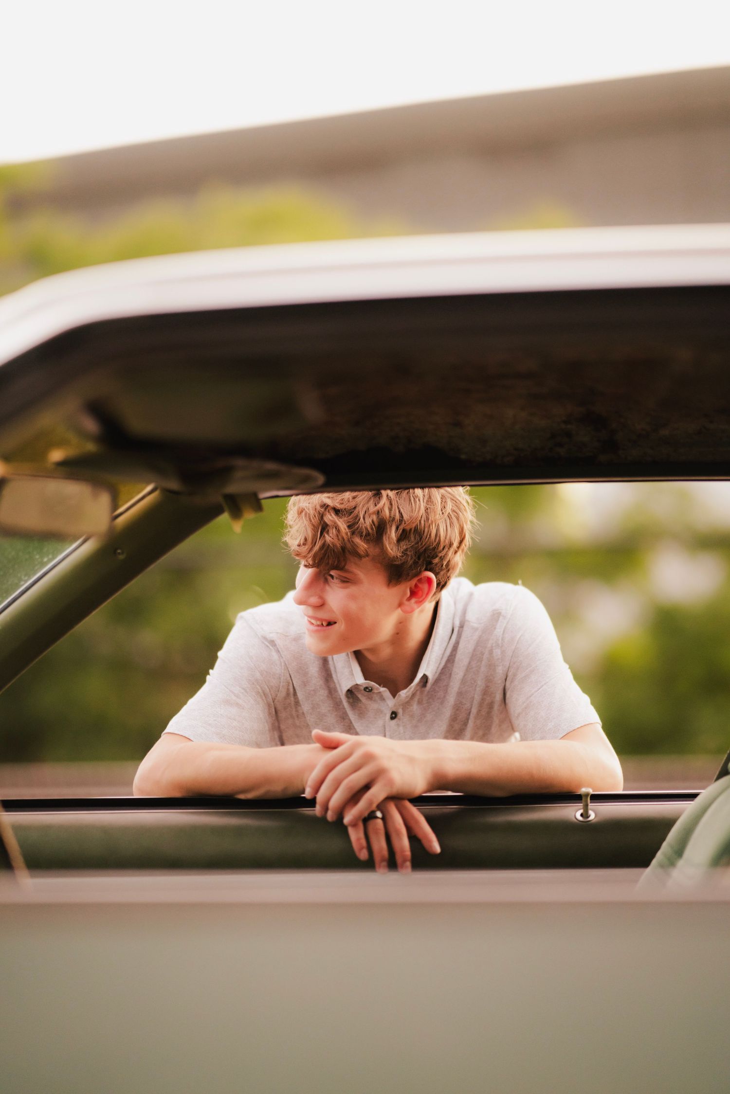 Young man leaning out of a vintage car window, smiling outdoors with green trees in the background.