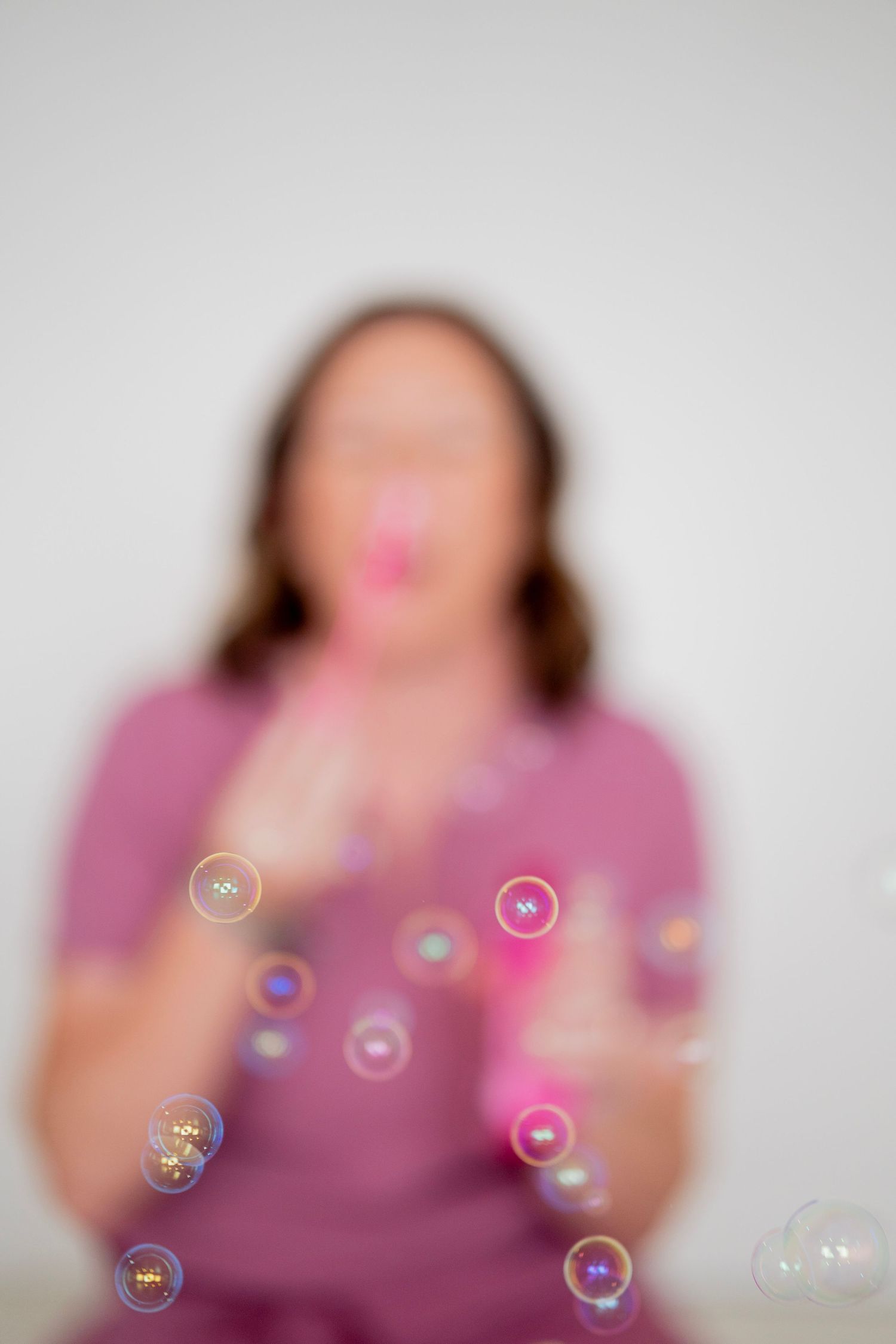 Woman in pink shirt blowing colorful soap bubbles against a white background, with bubbles in sharp focus.