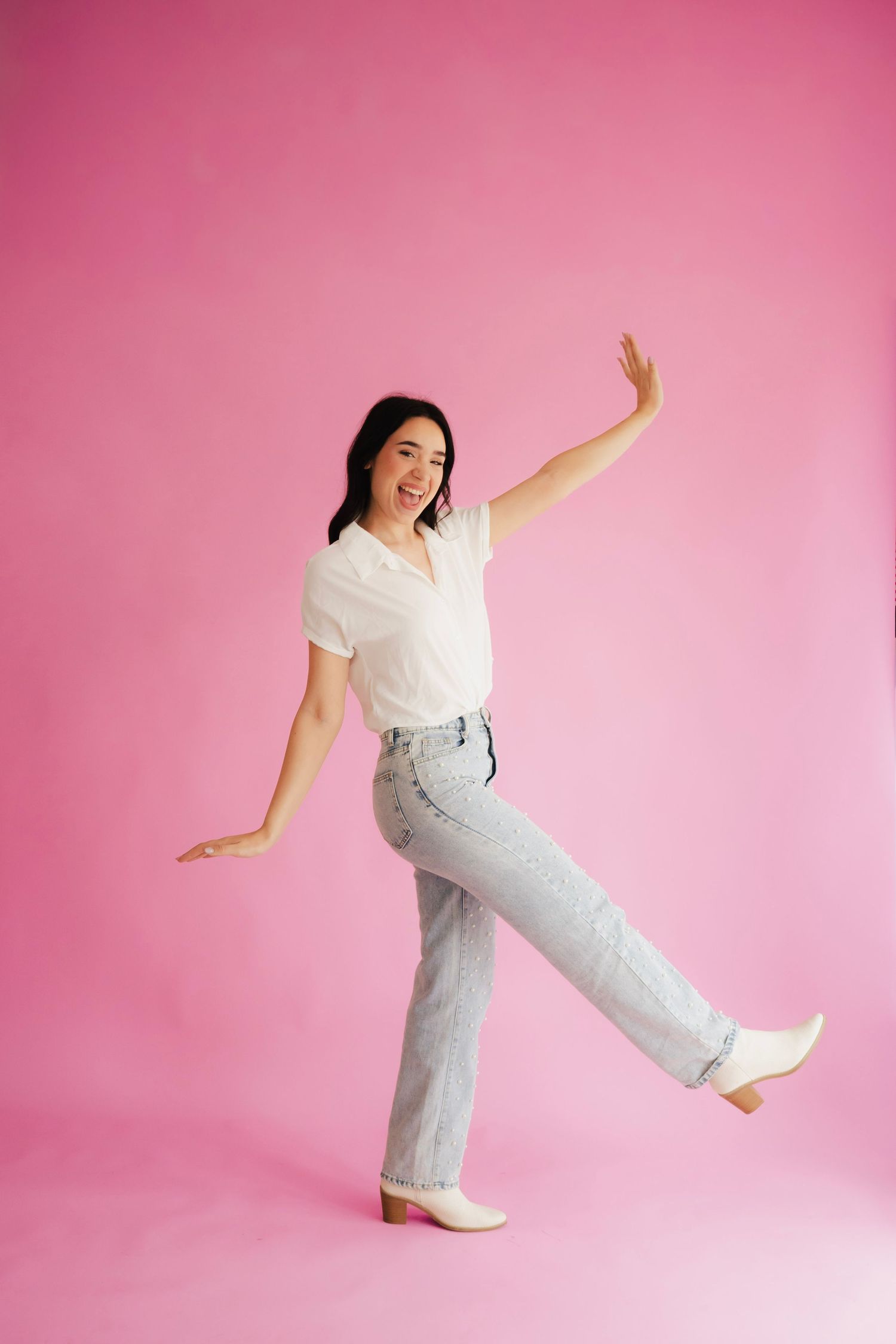 Young woman in white top and light jeans posing playfully on one leg against a pink background.
