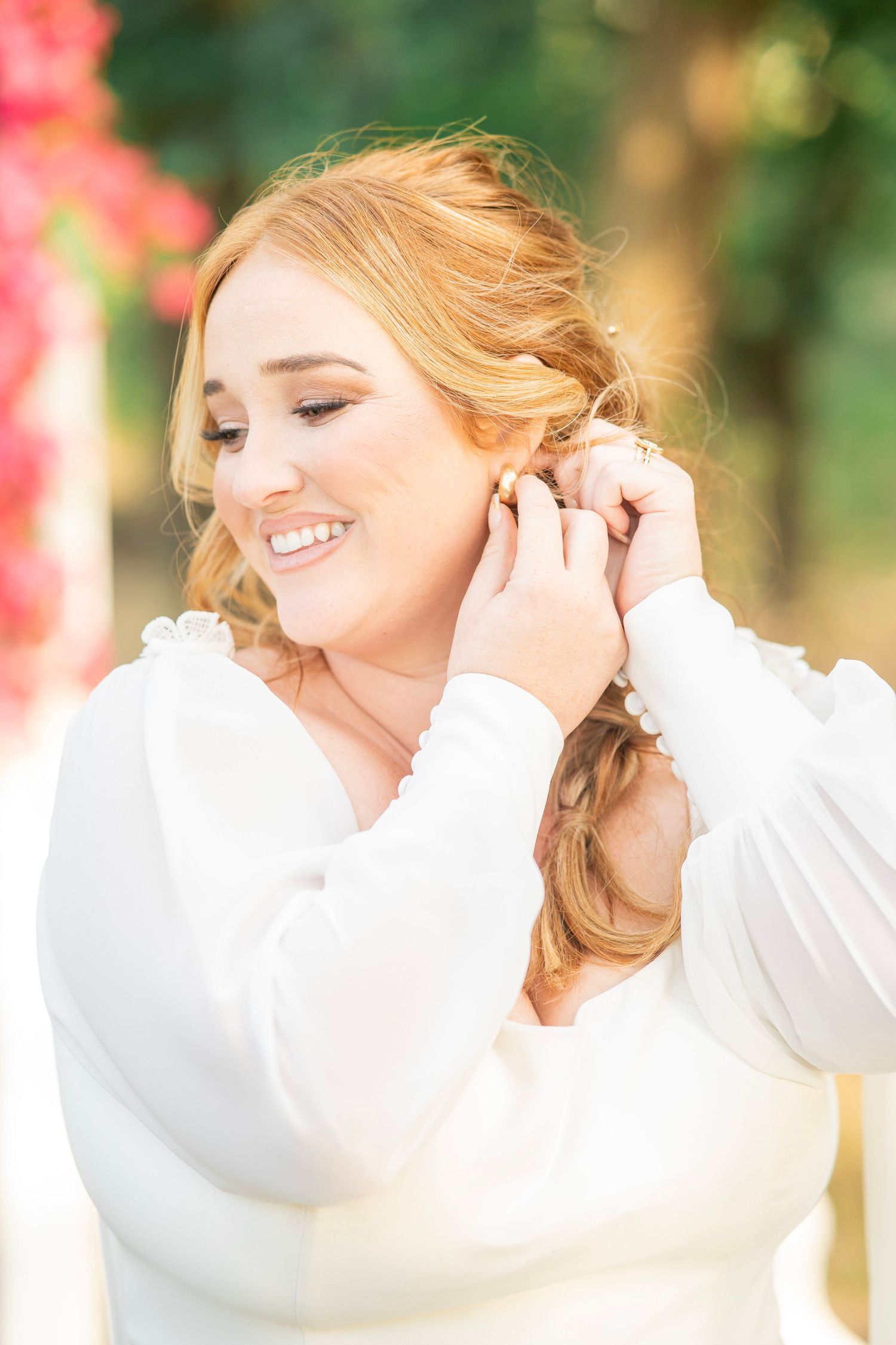 Smiling redhead bride putting on earrings outdoors before her wedding ceremony.