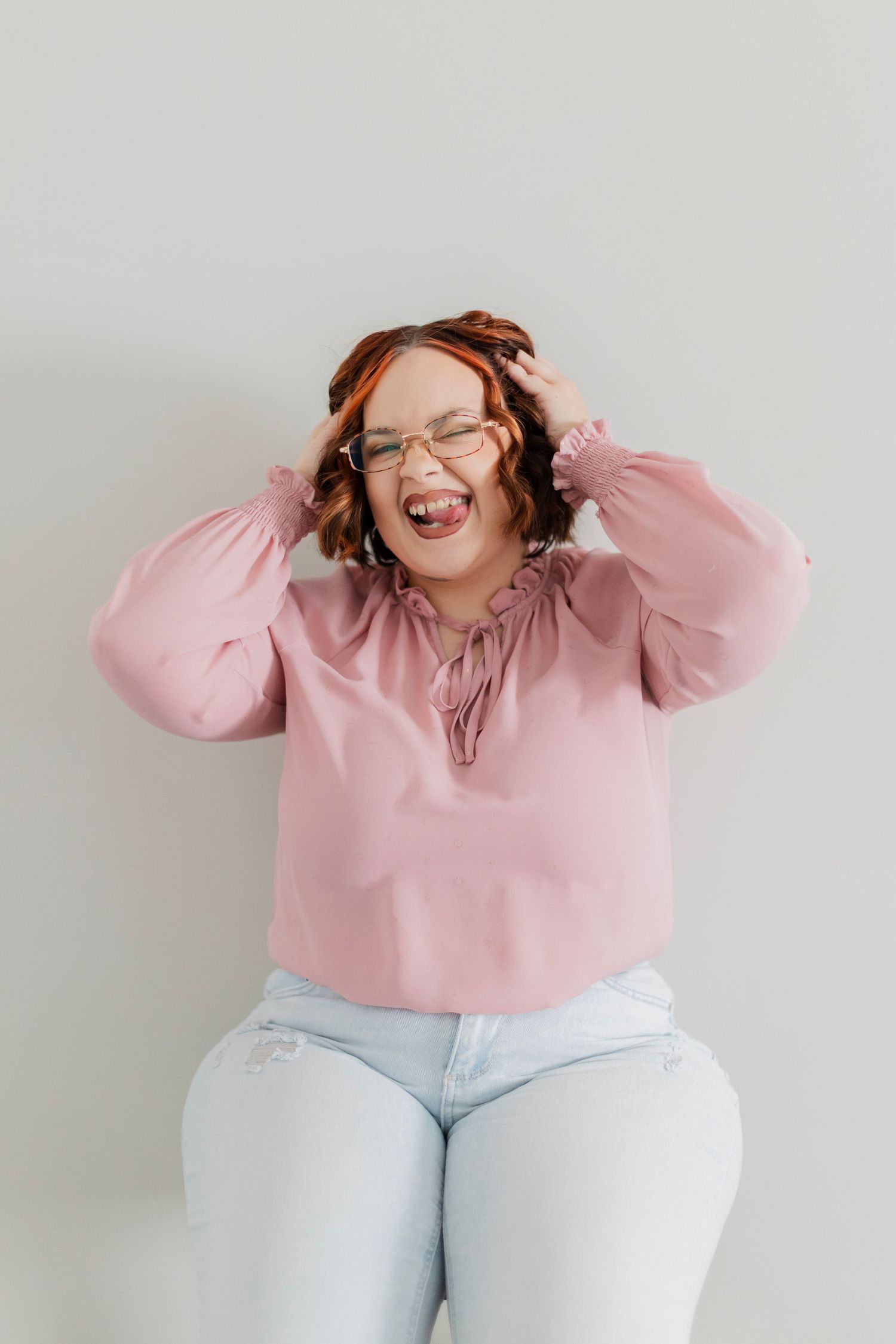 A joyful woman in a pink blouse and jeans laughs with hands in her hair against a light background.