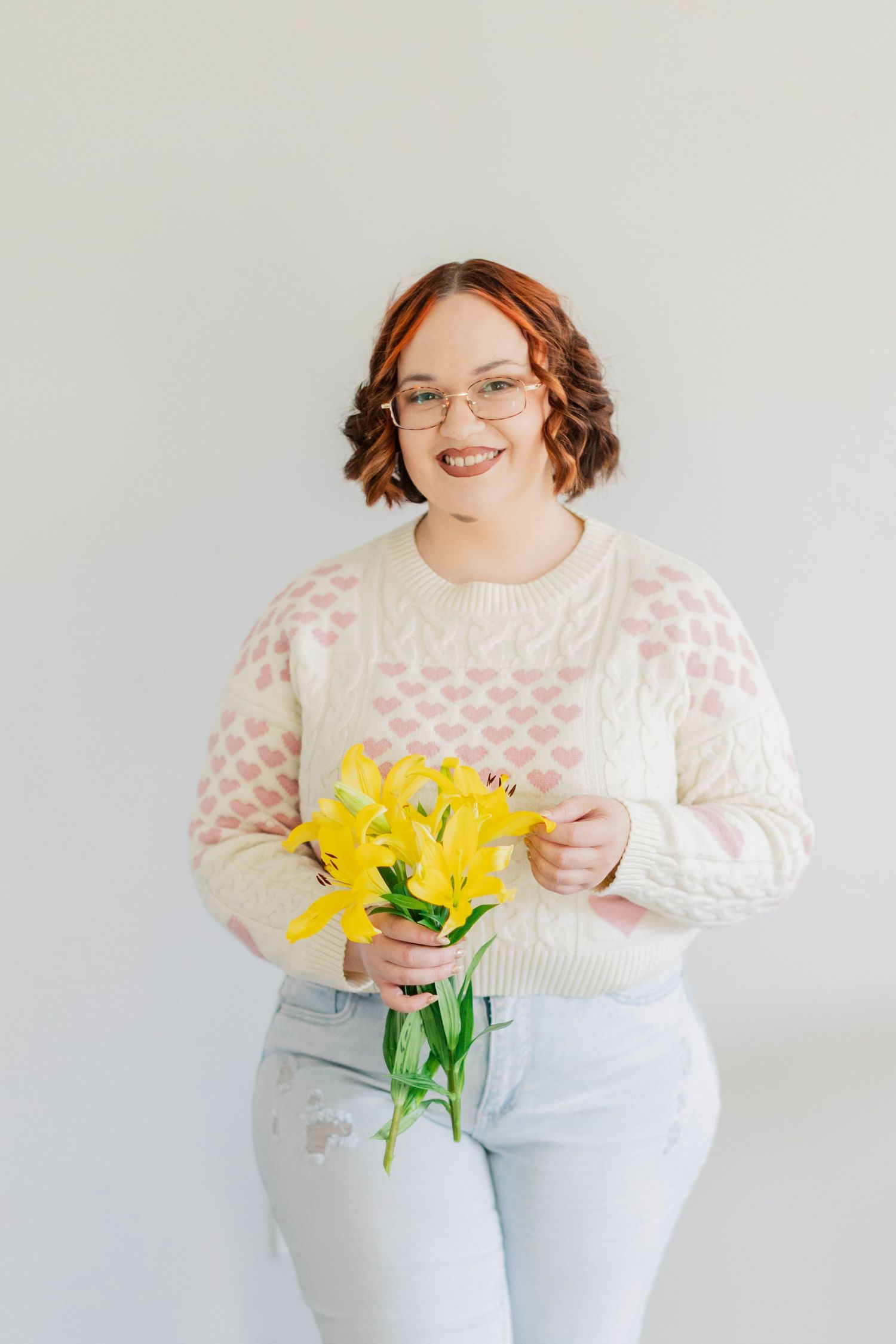 A smiling woman with red hair holds a bouquet of yellow lilies wearing a heart-print knit sweater.