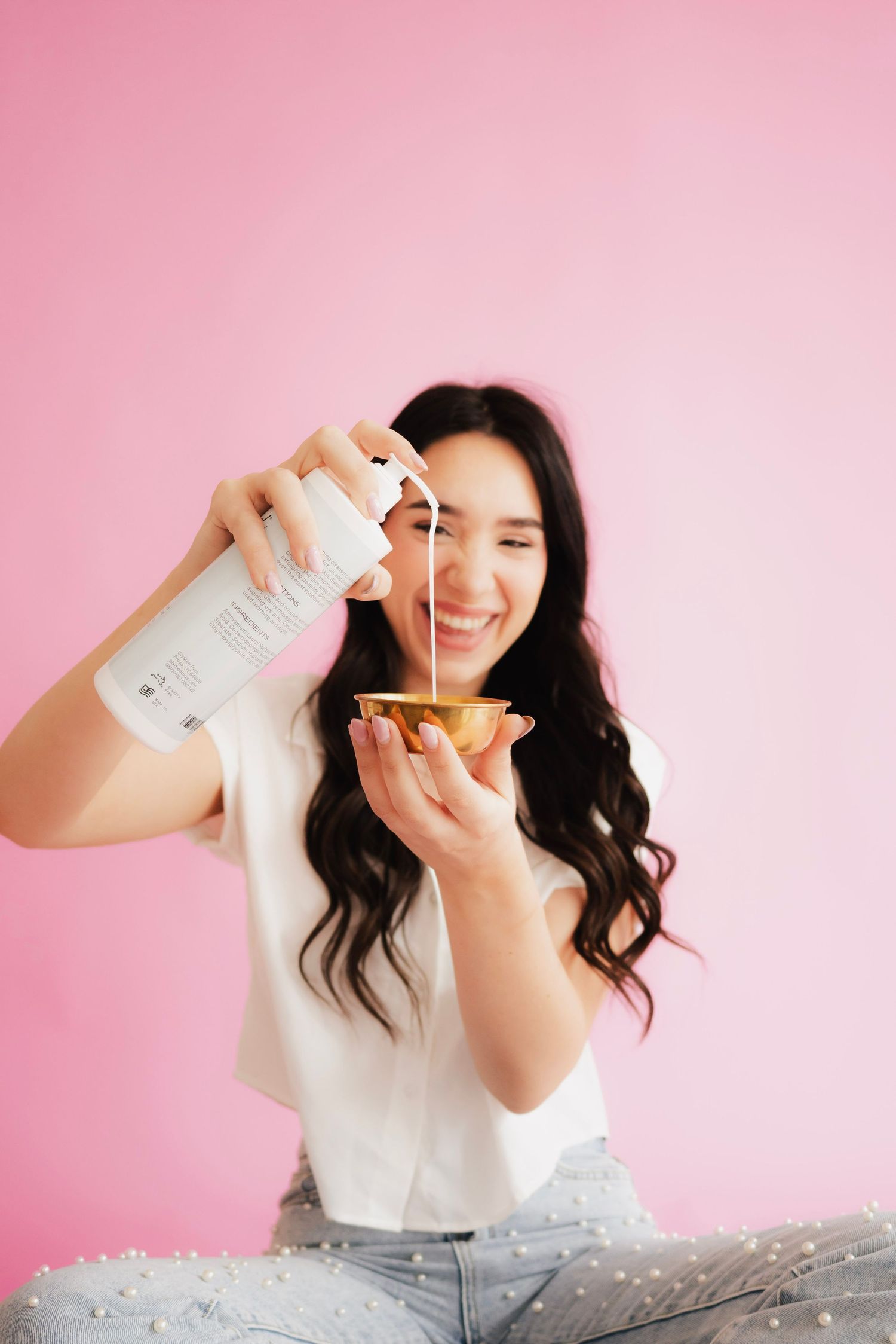Smiling woman pouring liquid from a spray bottle into a small gold bowl against a pink background.