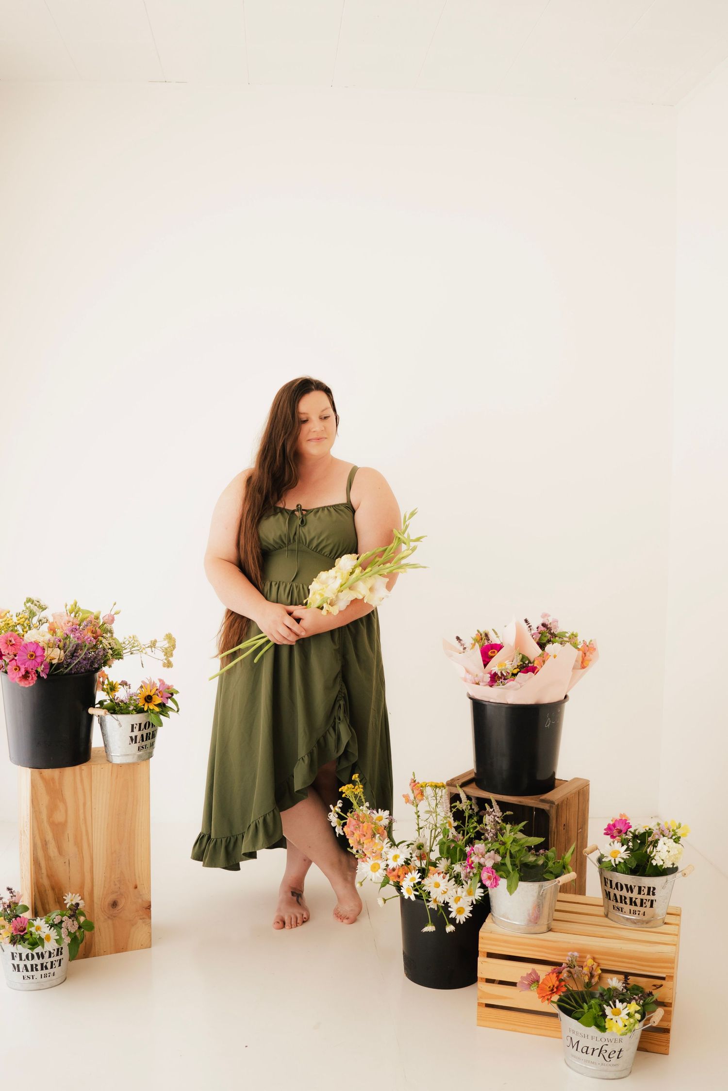 Woman in green dress holding flowers surrounded by buckets and crates of colorful blooms in a bright white studio.