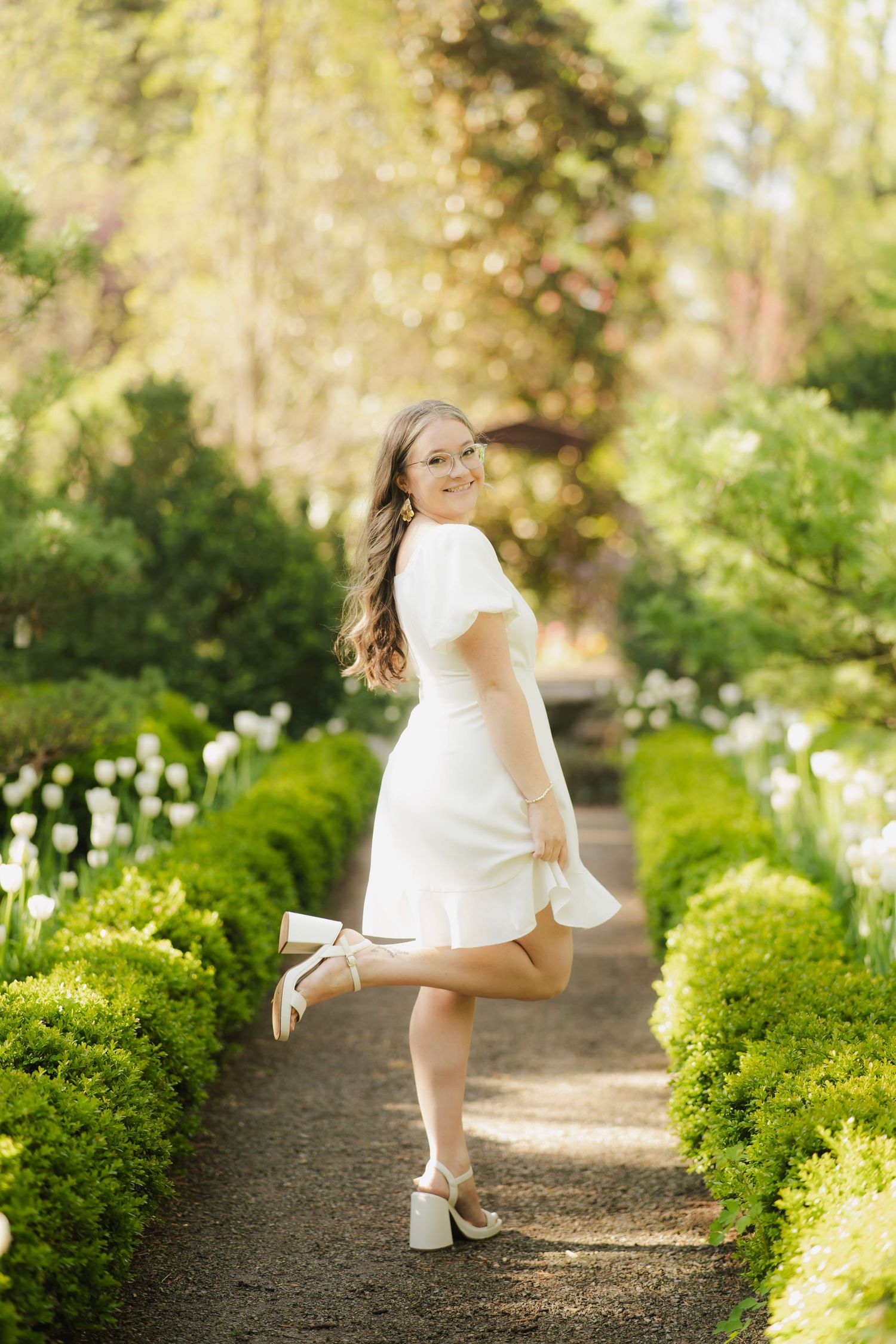 Happy young woman in white dress posing on garden path with manicured hedges.