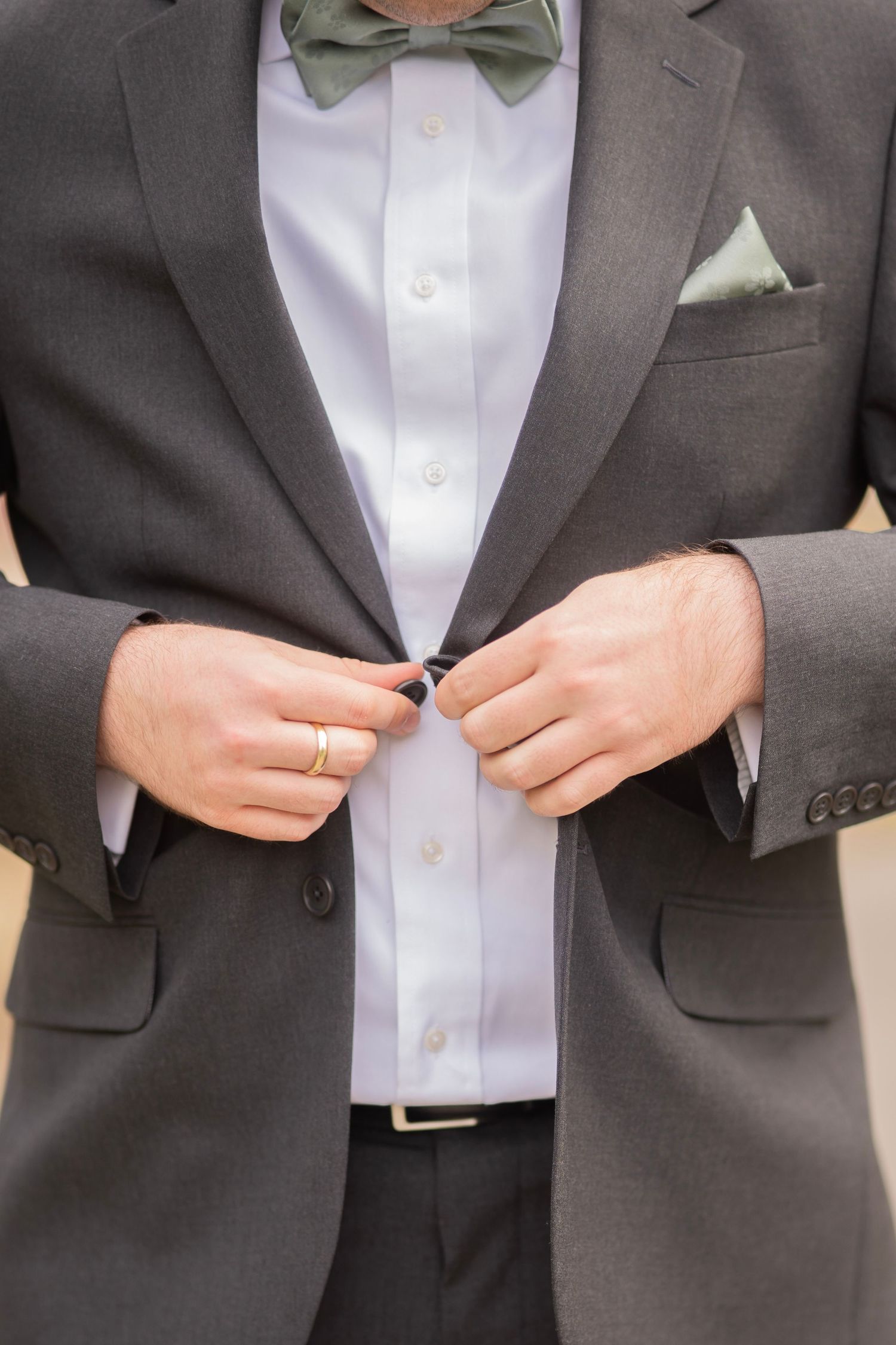 Groom in grey suit adjusting jacket button, wearing sage green bow tie and matching pocket square.