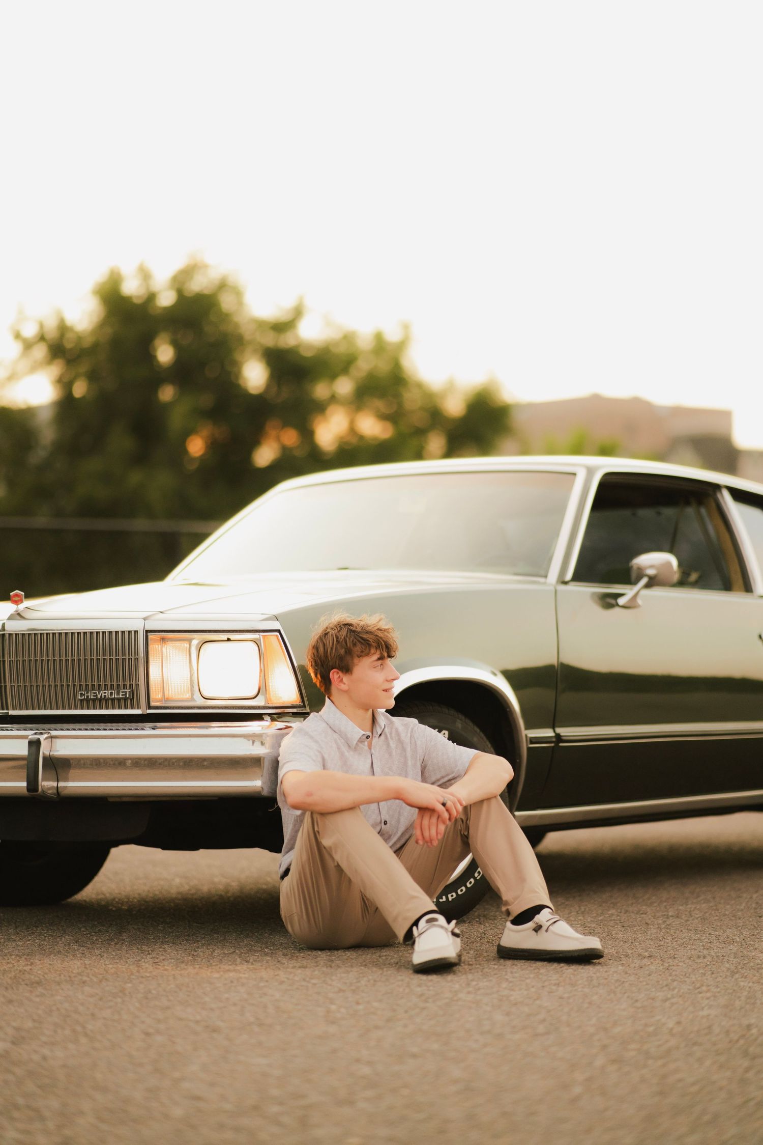Young man sitting on pavement leaning against a classic mint green car at golden hour.